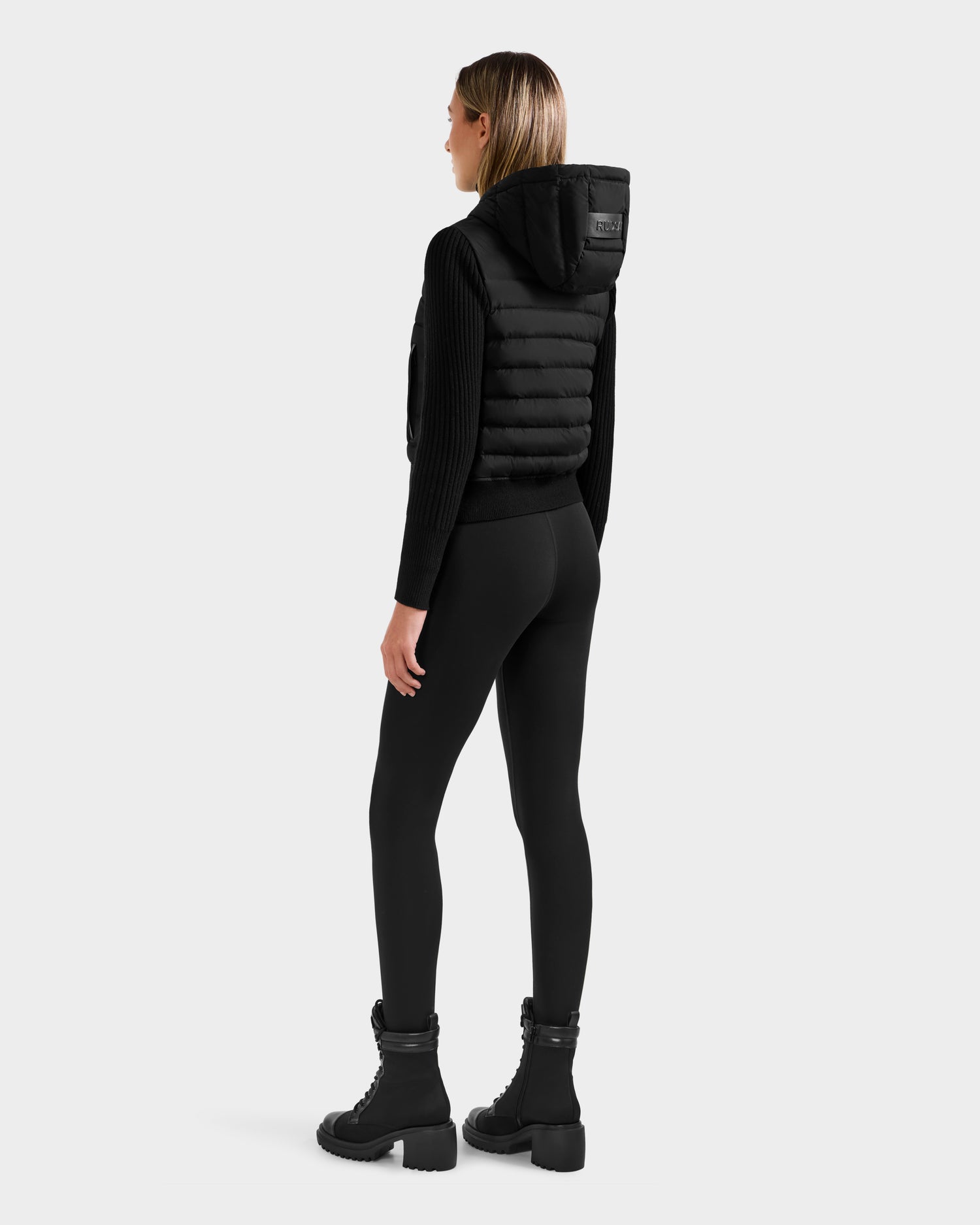 A woman stands facing away, wearing the Rudsak EMILY - L125515 Black hooded down bomber with RDS-certified down, a black ribbed sweater, black leggings, and chunky-heeled lace-up boots against a plain light gray background.