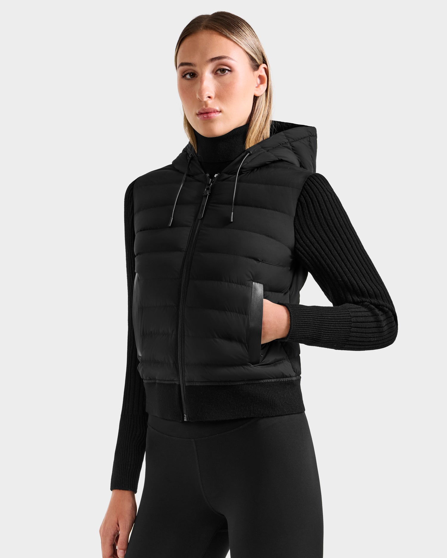 A woman wears the Rudsak EMILY - L125515 Black hooded down bomber vest over a black ribbed long-sleeve sweater and black leggings, standing against a plain light gray background.