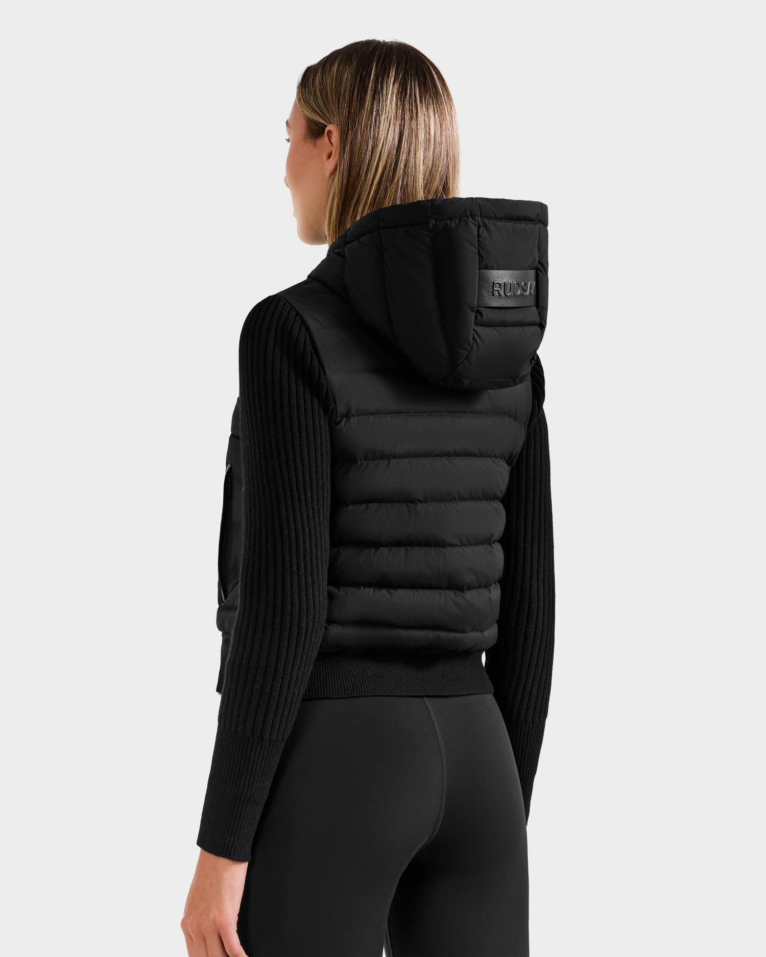 A woman with straight, shoulder-length hair models the Rudsak EMILY - L125515 Black puffer vest, featuring an RDS-certified down fill and hood, over a ribbed long-sleeve top and black leggings, facing away from a plain light background.
