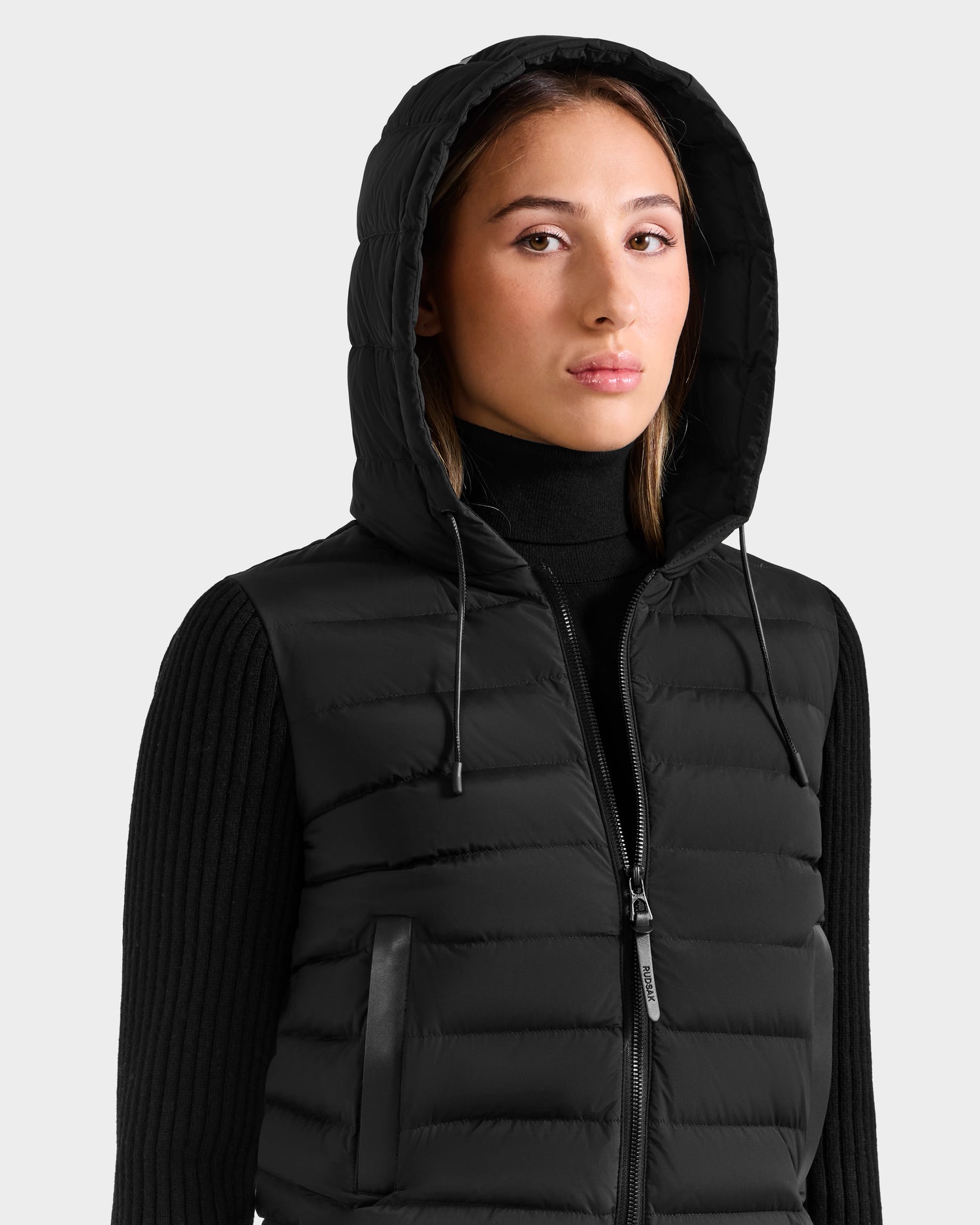 A person wears the EMILY - L125515 Black women’s hooded down bomber by Rudsak over a black ribbed turtleneck sweater, looking slightly to the side against a plain light background.