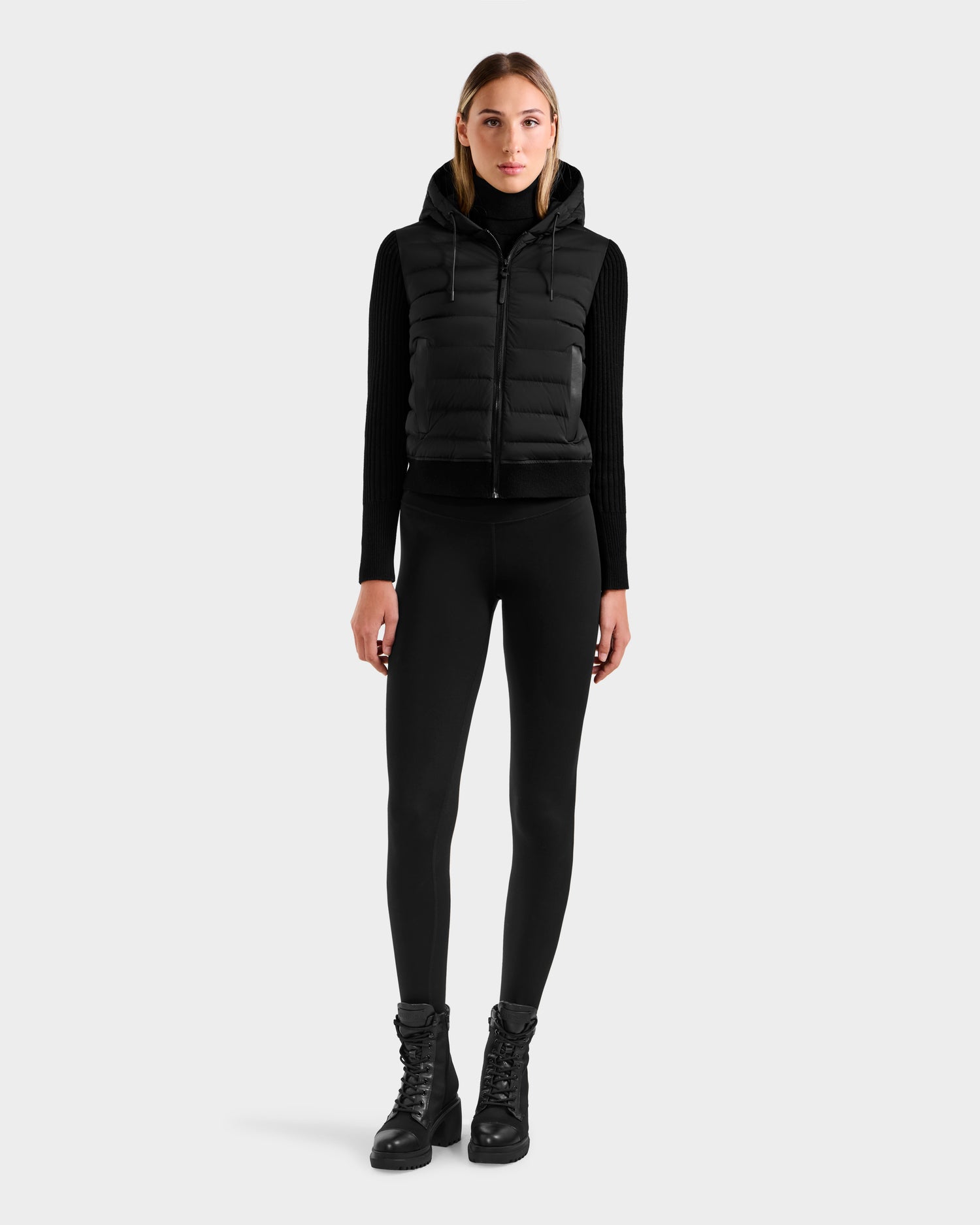 A woman faces forward against a plain light gray background, wearing the Rudsak EMILY - L125515 Black women’s hooded down bomber over a black long-sleeve top, black leggings, and black lace-up boots.