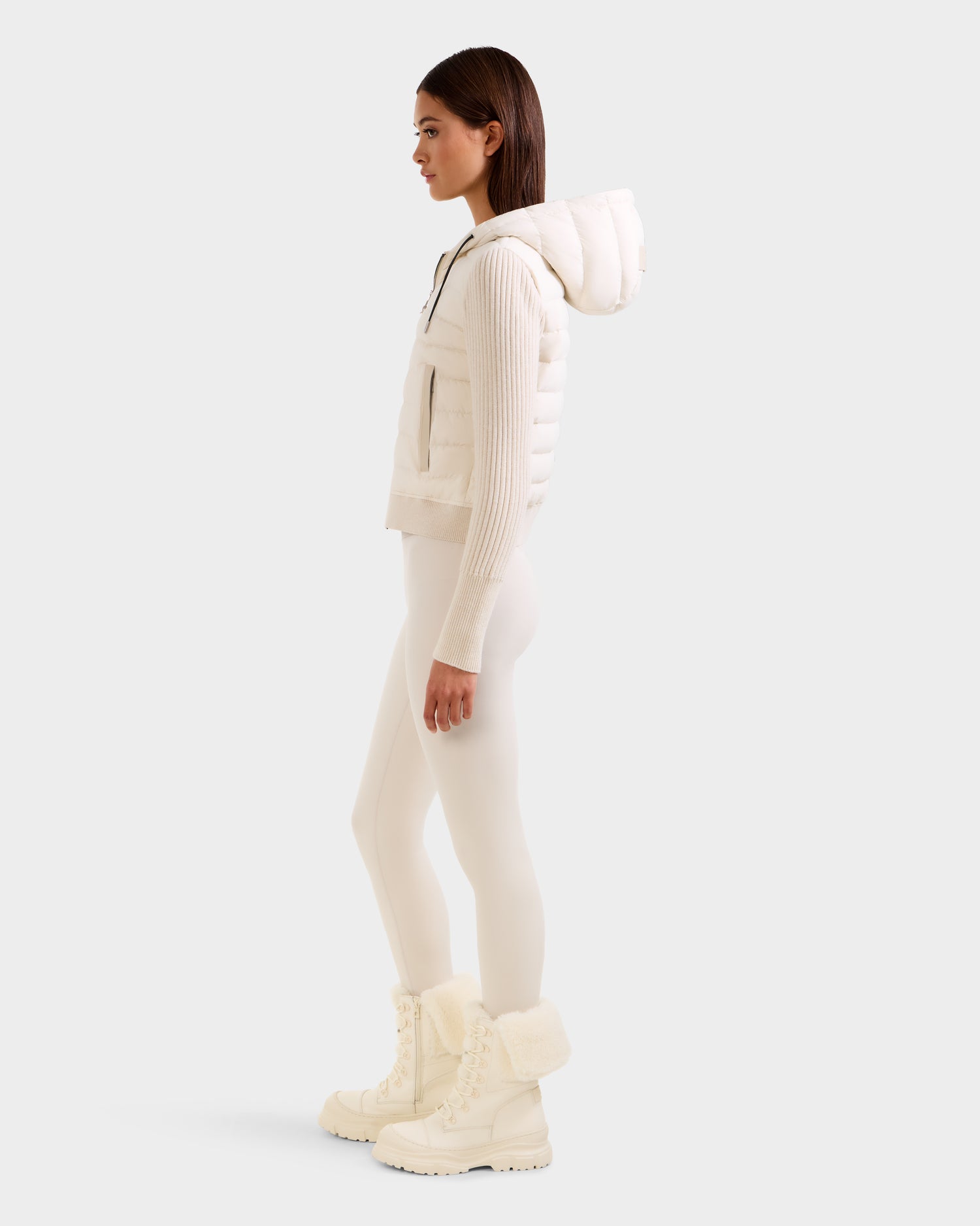A woman stands in profile against a plain light background wearing Rudsak’s EMILY - L125515 Vanilla, an all-white, modern cropped women's hooded down bomber. She pairs it with fitted leggings and fur-lined winter boots.
