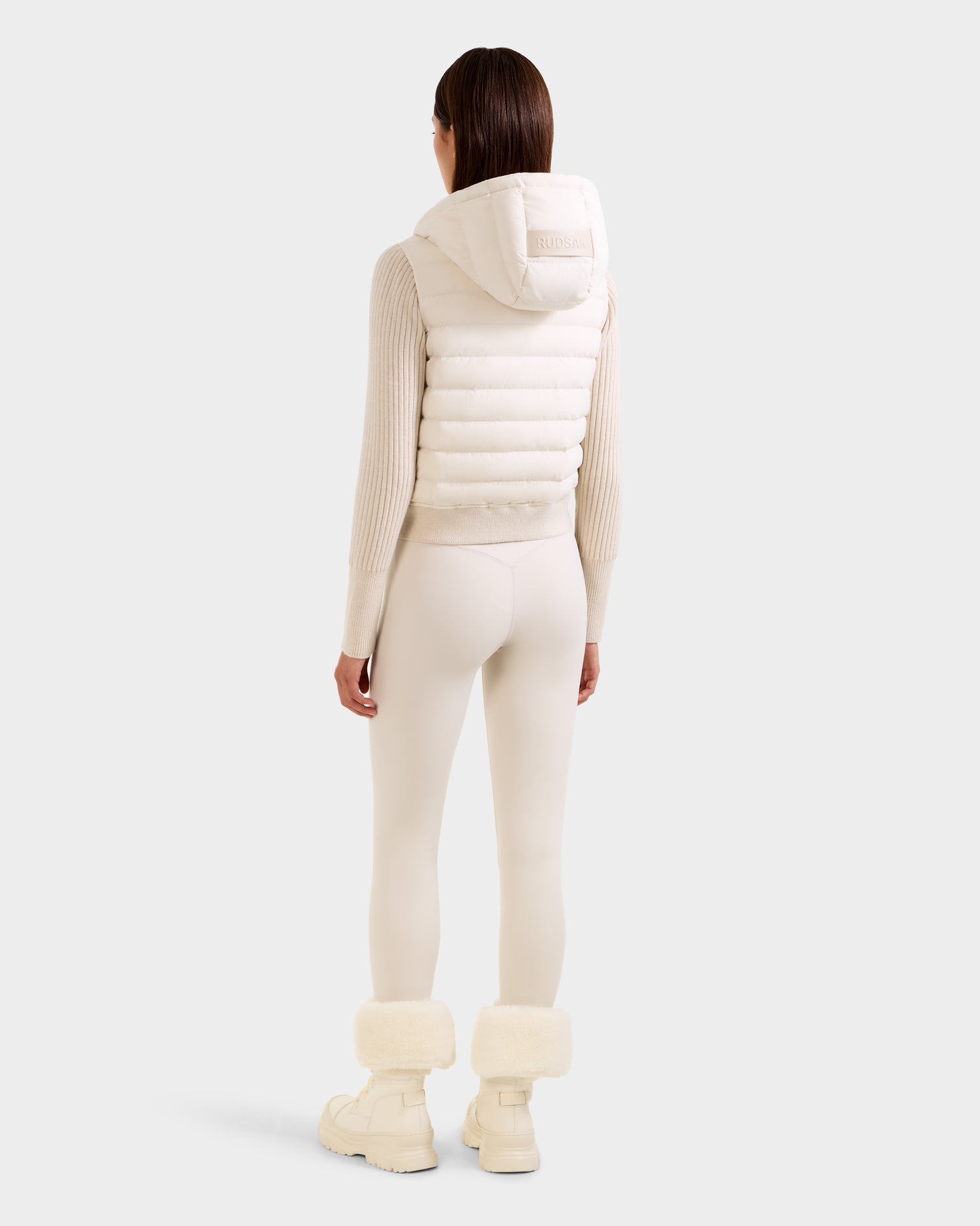 A person stands facing away, wearing the Rudsak EMILY - L125515 Vanilla women’s hooded down bomber with a cropped silhouette, paired with cream ribbed top, fitted leggings, and fur-trimmed boots against a light background.