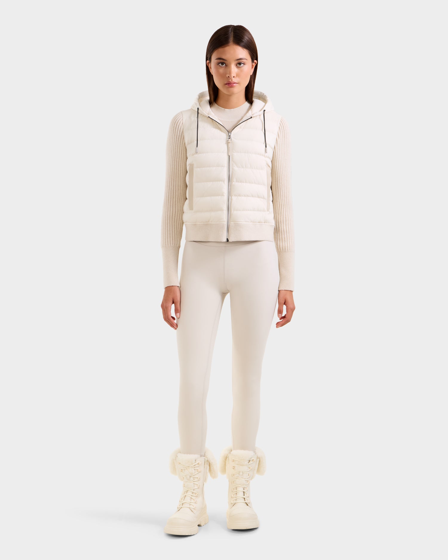 A woman faces forward in the Rudsak EMILY - L125515 Vanilla, a white RDS-certified down hooded puffer vest with a cropped modern silhouette, layered over a ribbed sweater, white leggings, and white lace-up fur-trimmed boots, against a light background.