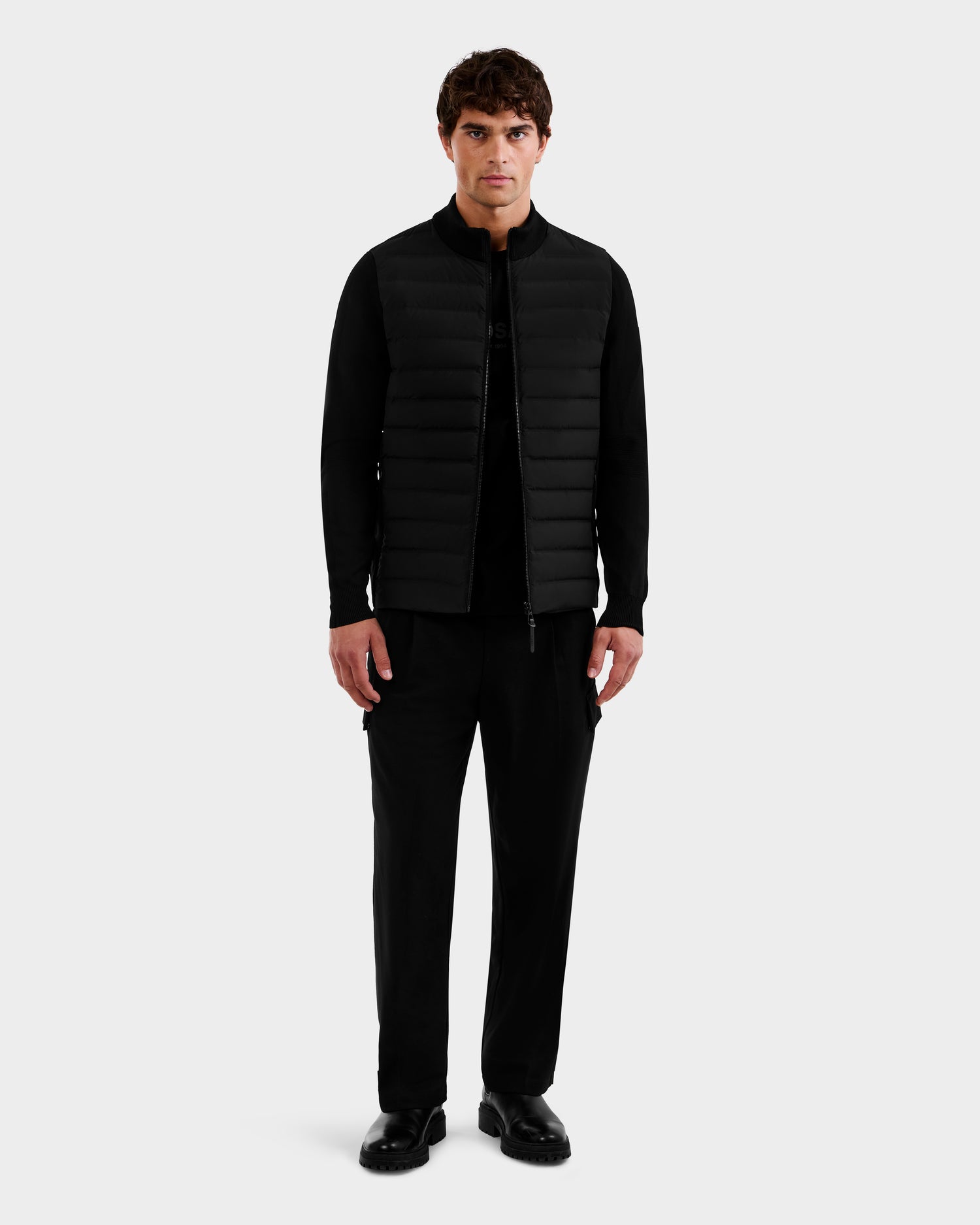 A man stands facing forward in Rudsak’s EMMETT - 6125029 Black mock-neck hybrid jacket with lightweight down insulation, paired with black shirt, pants, and shoes against a plain light gray background—a wardrobe staple.