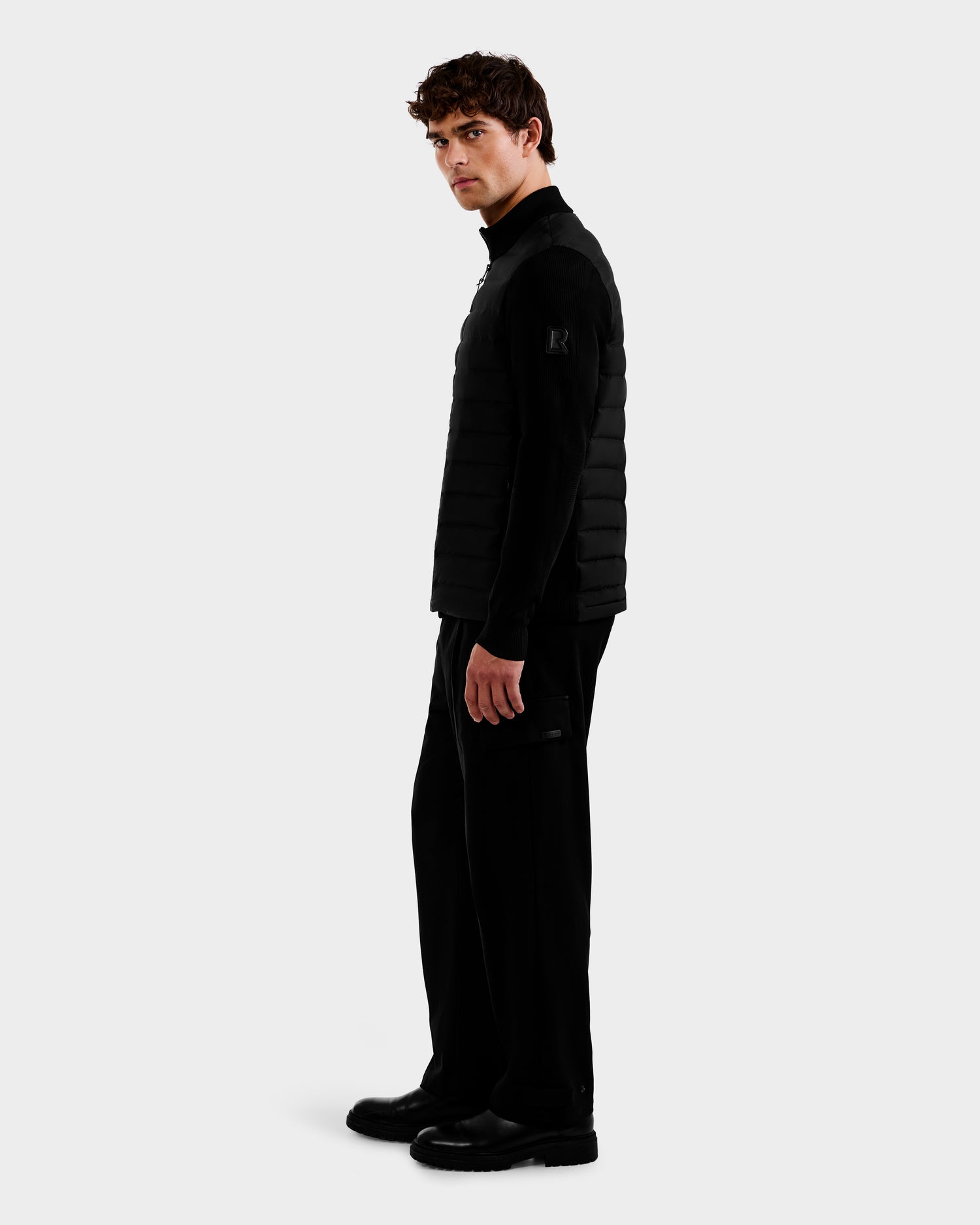 A man with curly brown hair models the Rudsak EMMETT - 6125029 Black mock-neck hybrid jacket, featuring lightweight down insulation, paired with black pants and shoes, standing sideways against a plain white background.