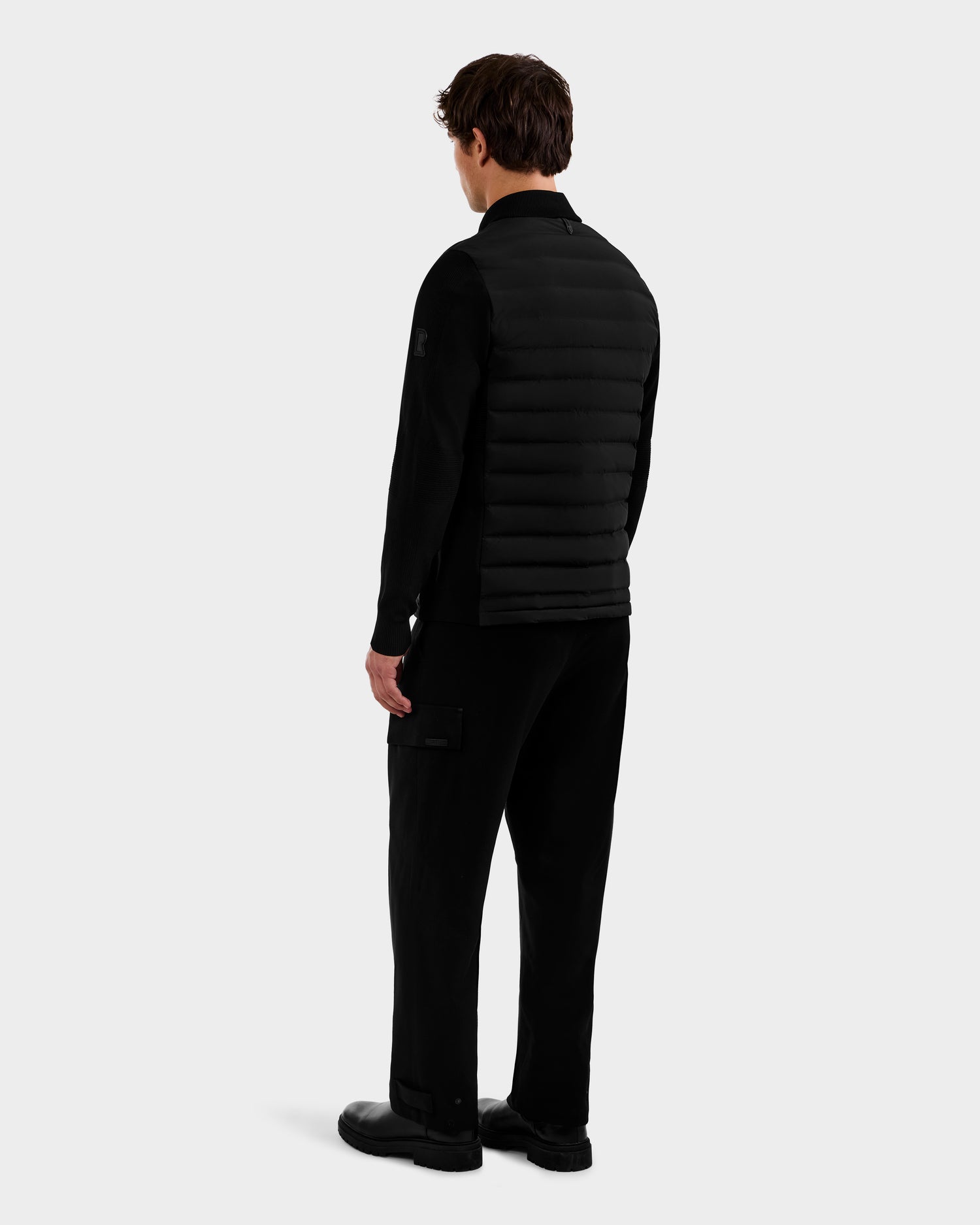 A man faces away, wearing the Rudsak EMMETT - 6125029 Black mock-neck hybrid jacket with lightweight down insulation, paired with black cargo pants and shoes, set against a plain light gray background—modern menswear essentials.
