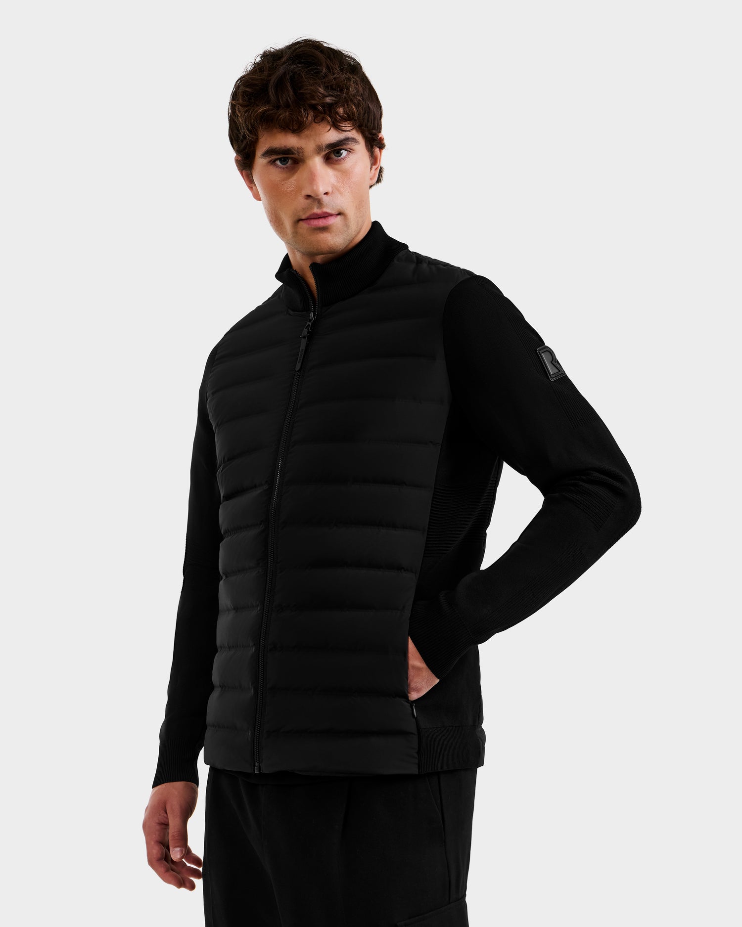 A man with short brown hair models the Rudsak EMMETT - 6125029 Black mock-neck hybrid jacket and matching black pants, standing against a plain light gray background with one hand in his pocket.