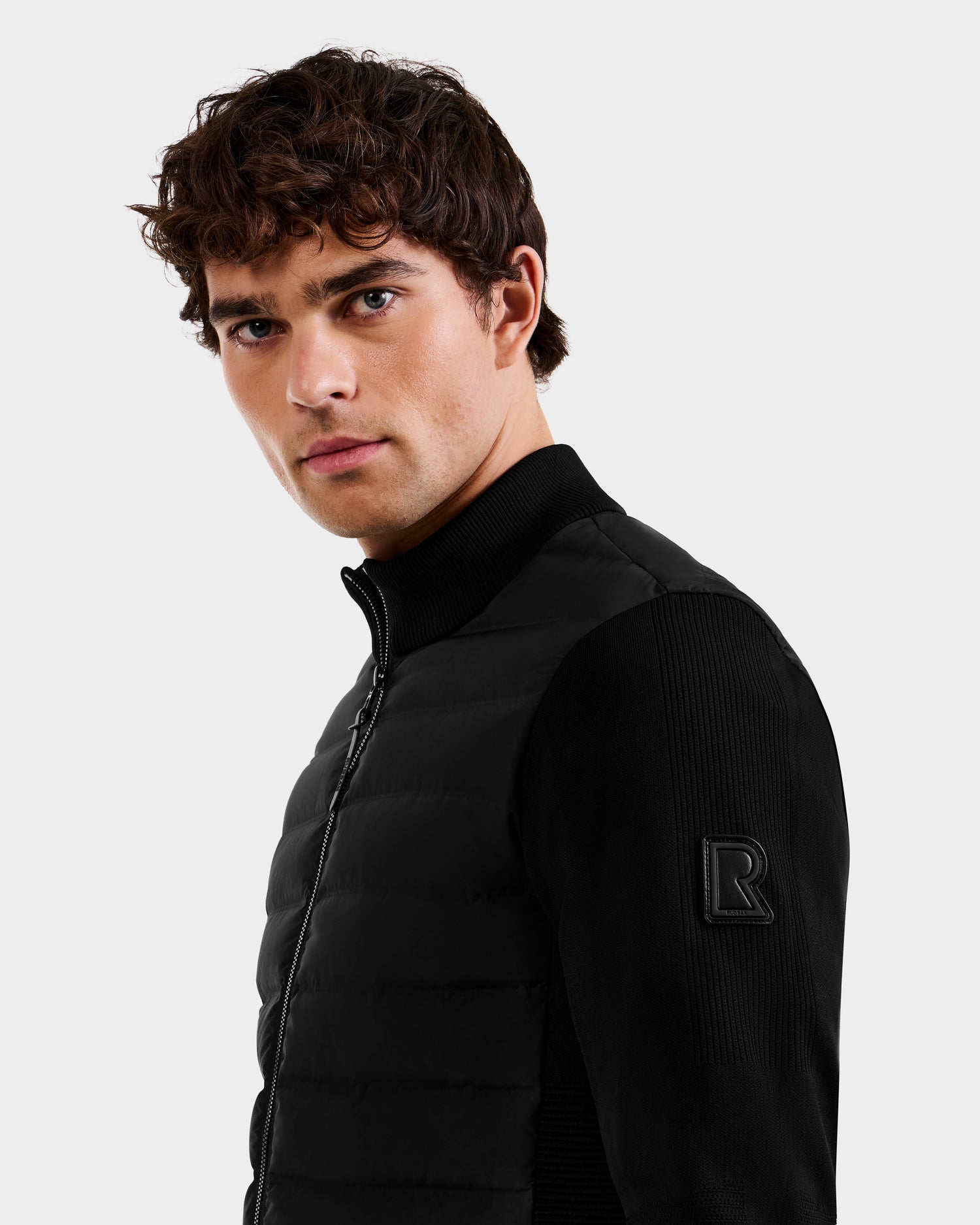 A young man with curly brown hair wears the Rudsak EMMETT - 6125029 Black mock-neck hybrid jacket, featuring ribbed sleeves and an embossed R on the shoulder, posing against a light gray background—perfect for the modern men's wardrobe.