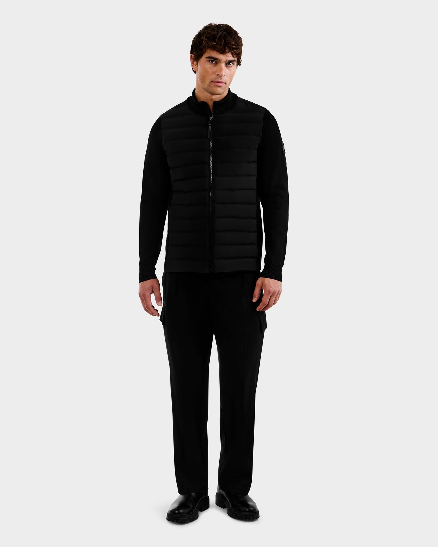 A man stands facing forward, wearing the Rudsak EMMETT - 6125029 Black hybrid jacket with horizontal quilting and lightweight down insulation, paired with black pants and shoes, against a plain light gray background.