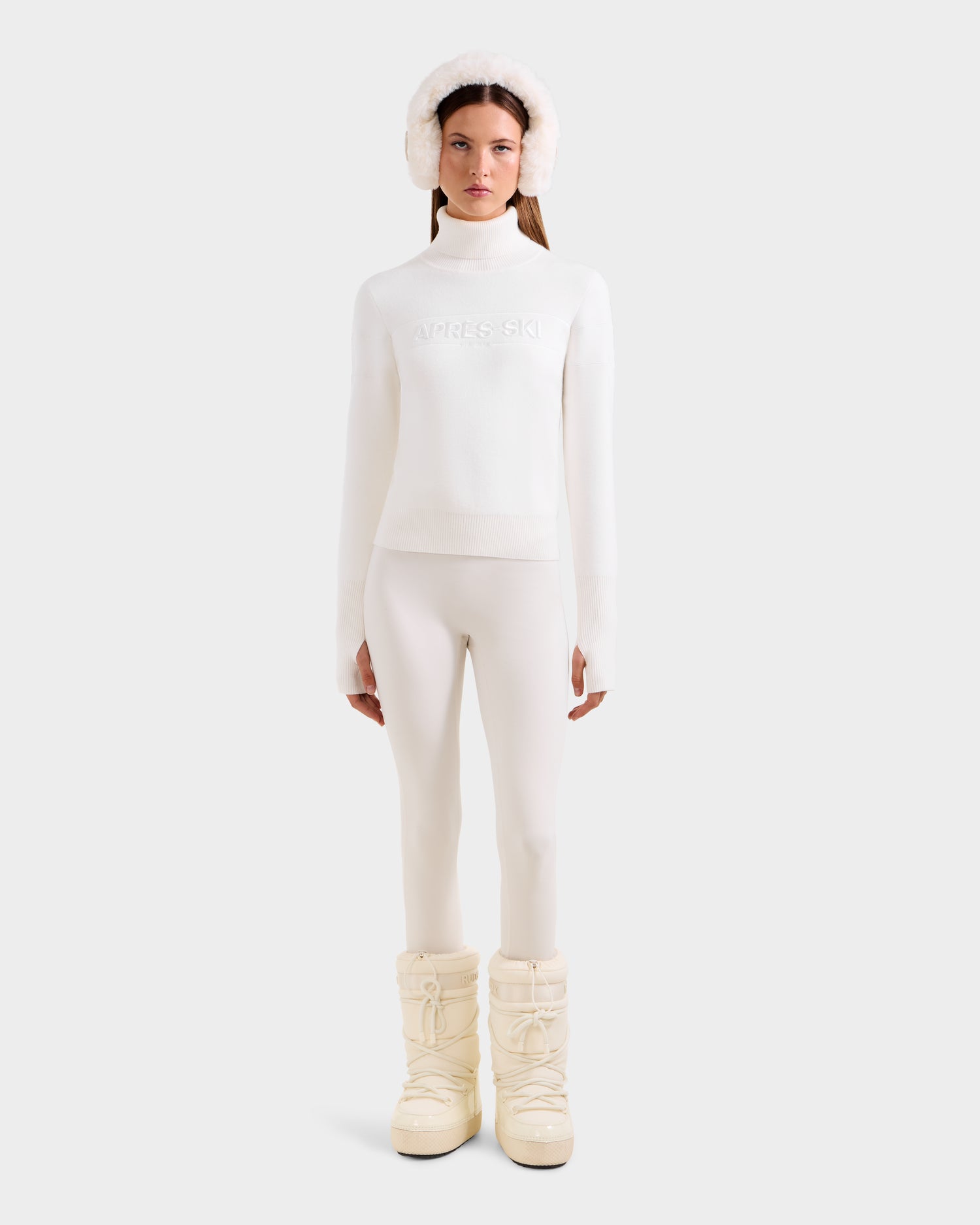 A woman stands facing forward, wearing the Rudsak ERIKA - 8523536 Iceland turtleneck, fitted pants, fluffy earmuffs, and chunky snow boots—an effortlessly chic winter look against a plain background.