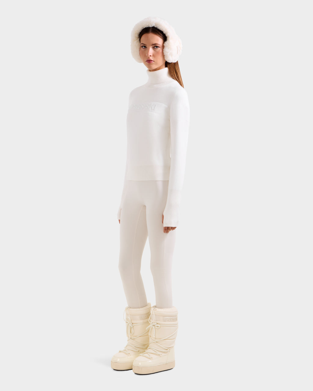 A woman faces forward in a chic winter look, wearing the Rudsak ERIKA - 8523536 Iceland turtleneck sweater, leggings, fluffy earmuffs, and tall padded snow boots against a plain light gray background.