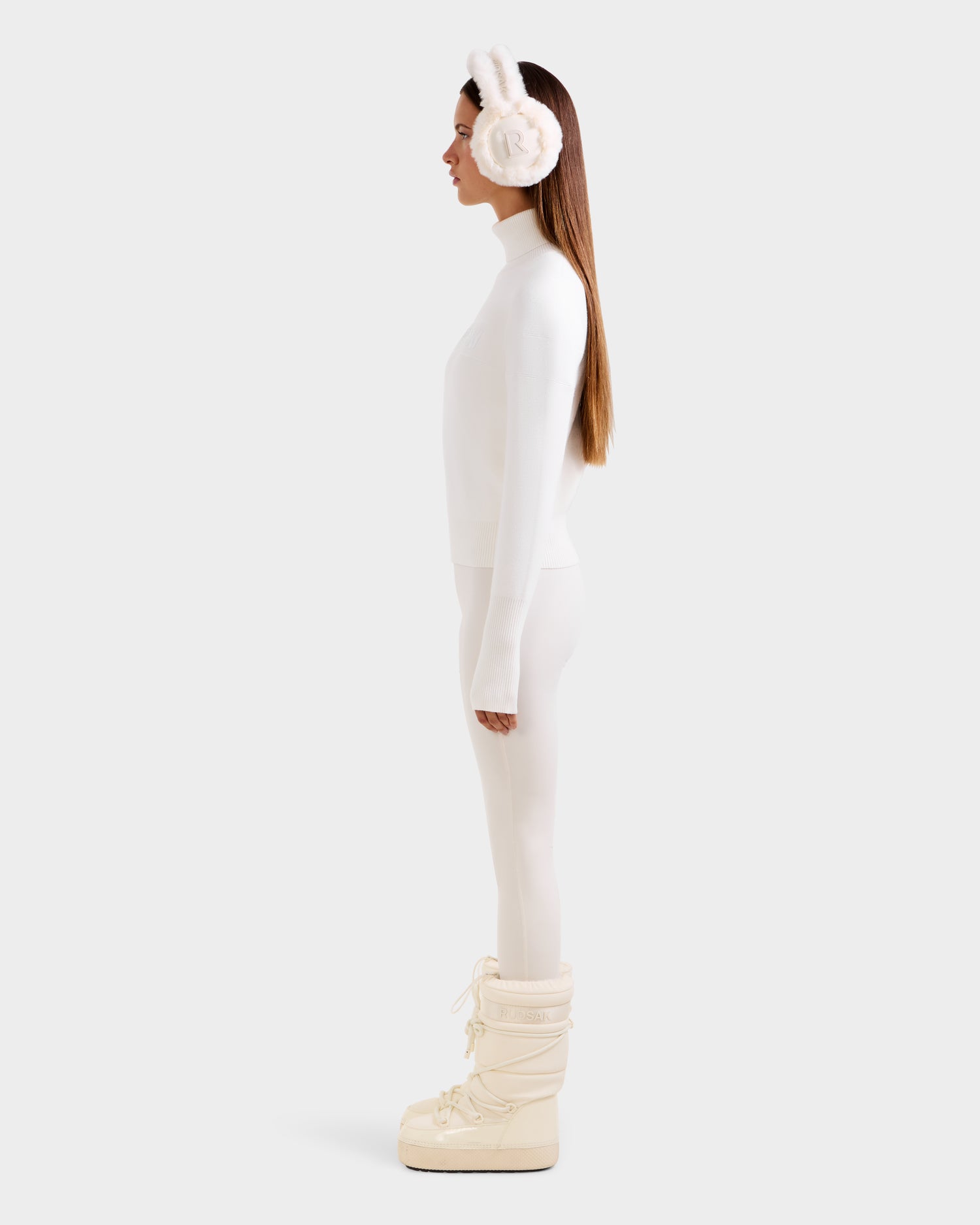 A woman stands in profile against a white background, wearing the Rudsak ERIKA - 8523536 Iceland turtleneck sweater with white bunny ear earmuffs, white leggings, and cream winter boots for a chic winter look.