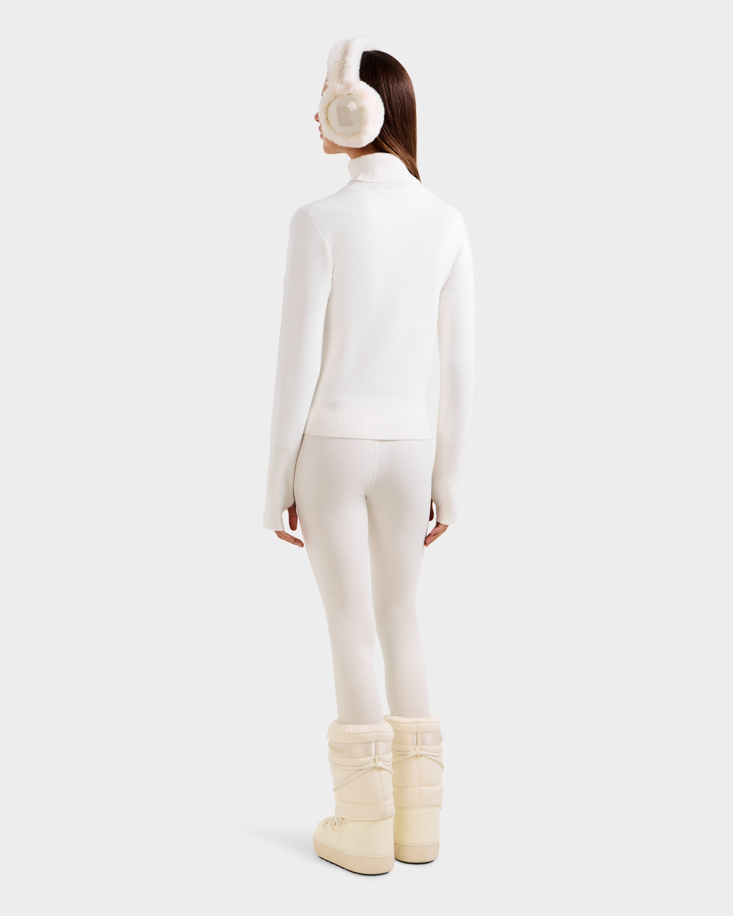 A person stands facing away, wearing Rudsak ERIKA - 8523536 Iceland earmuffs, an après-ski turtleneck sweater, white leggings, and beige winter boots against a light background—chic essentials for a winter wardrobe.