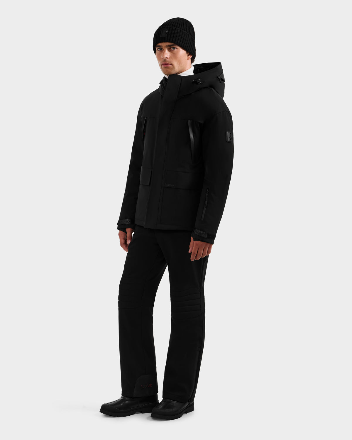 A person stands against a plain background wearing the Rudsak ETHAN - 6125589 Black men’s ski jacket, paired with a black beanie, pants, and boots, looking slightly to the side with a neutral expression.