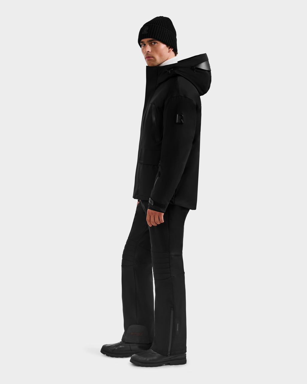 A person wearing Rudsak’s ETHAN - 6125589 Black men’s ski jacket, black beanie, pants, and boots stands facing left, glancing at the camera against a plain light gray background.