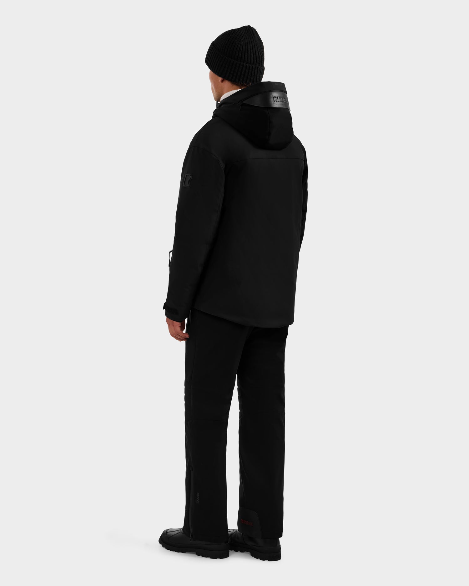 A person faces away, wearing the Rudsak ETHAN - 6125589 Black men’s ski jacket with RDS-certified down fill, paired with black pants and boots, set against a plain light gray background.