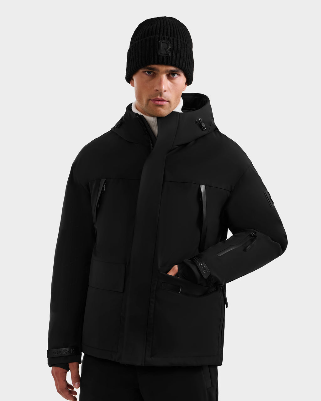 A man wearing the Rudsak ETHAN - 6125589 Black men’s ski jacket, filled with RDS-certified down and featuring multiple pockets, stands against a light gray background with one hand in his pocket.