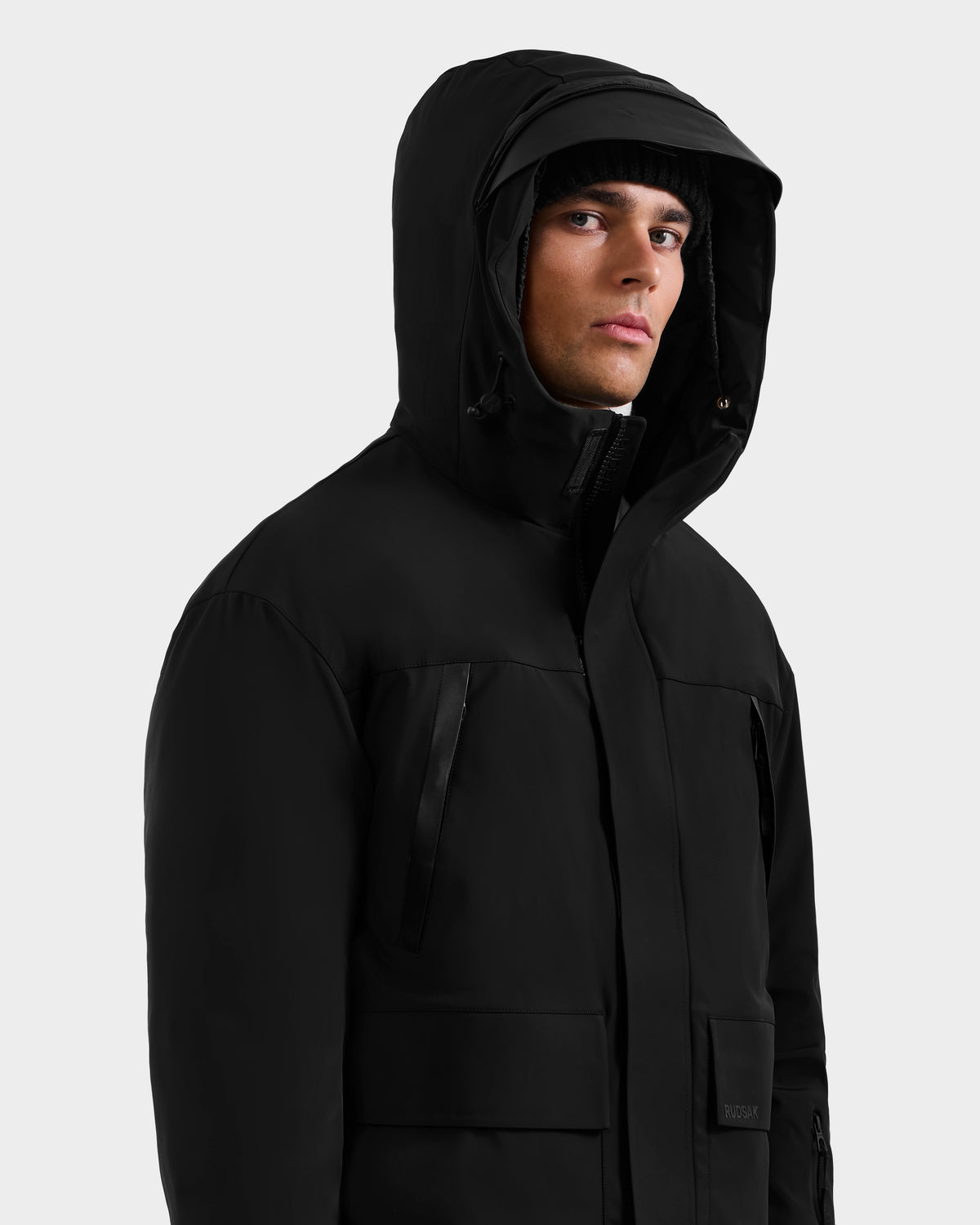 A man wearing the Rudsak ETHAN - 6125589 Black down jacket and a black beanie looks slightly to the side against a plain light gray background.