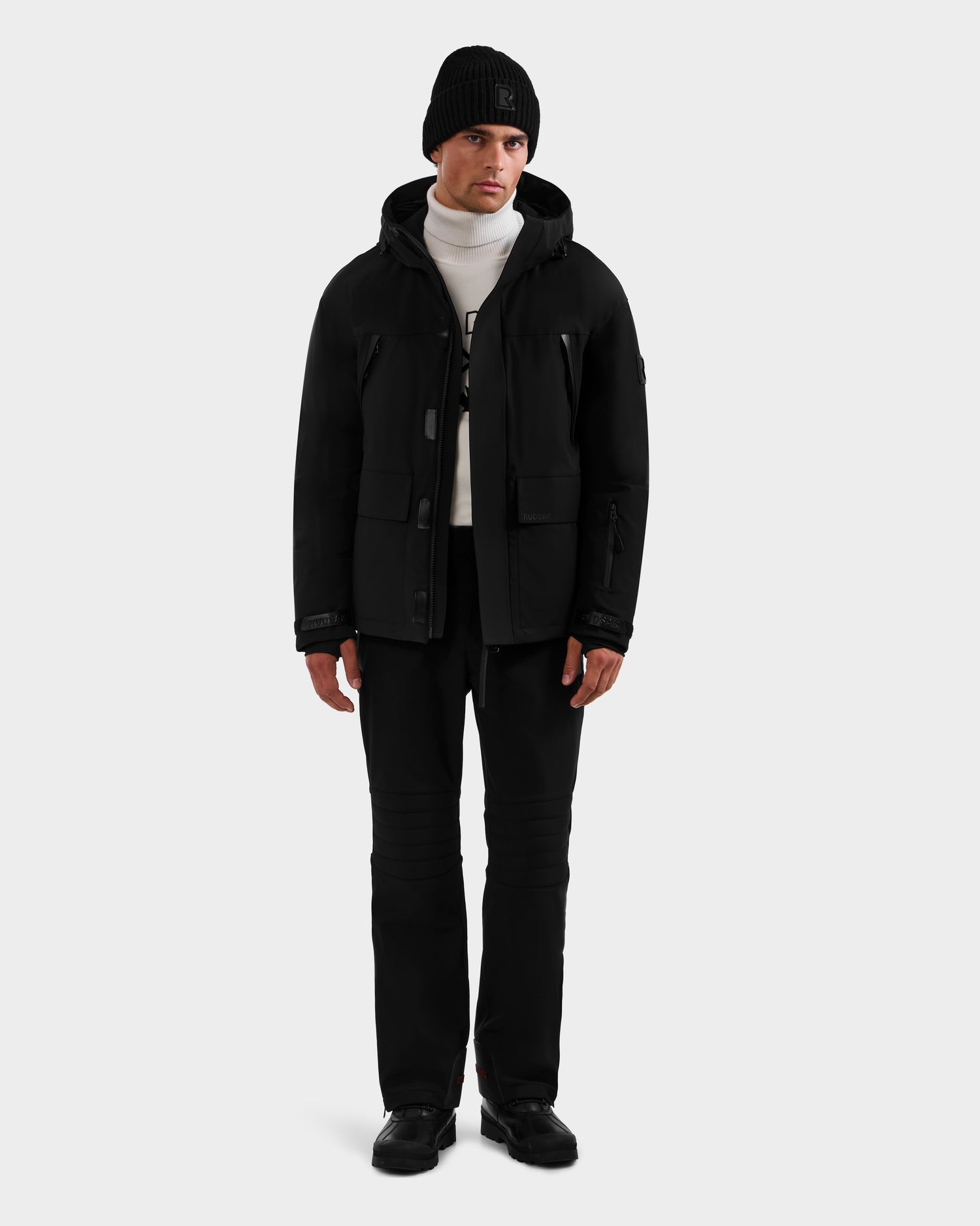 A man faces forward with a neutral expression, wearing the Rudsak ETHAN - 6125589 Black men’s ski jacket, black beanie, white turtleneck sweater, black pants, and black shoes against a plain background.