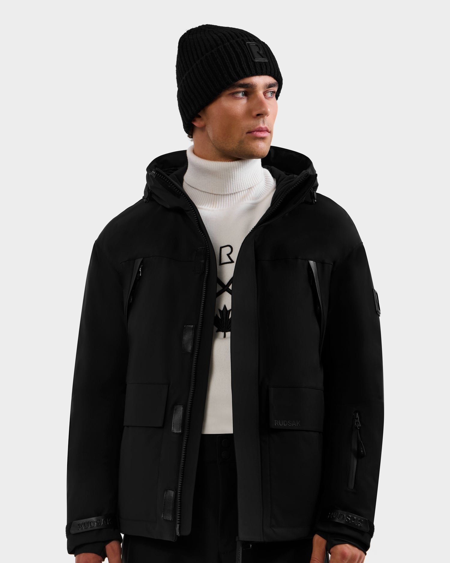 A man wearing the Rudsak ETHAN - 6125589 Black men’s ski jacket, a black knit beanie, a white turtleneck sweater with a black design, and black pants stands against a plain light background, looking slightly to his right.