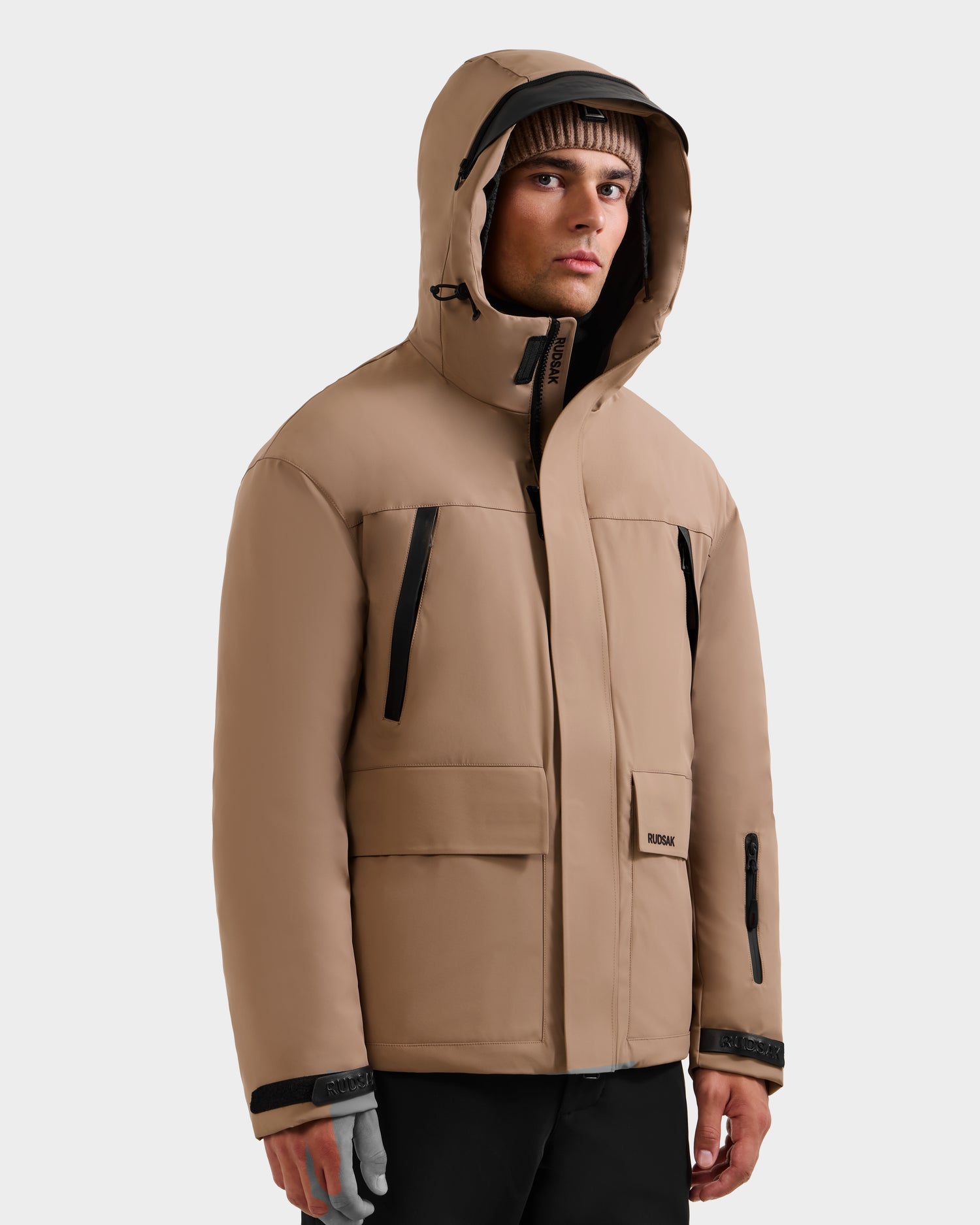 A man in the Rudsak ETHAN - 6125589 Sand ski jacket with black zippers and accents stands facing slightly left, paired with a brown beanie and black pants against a plain light background.