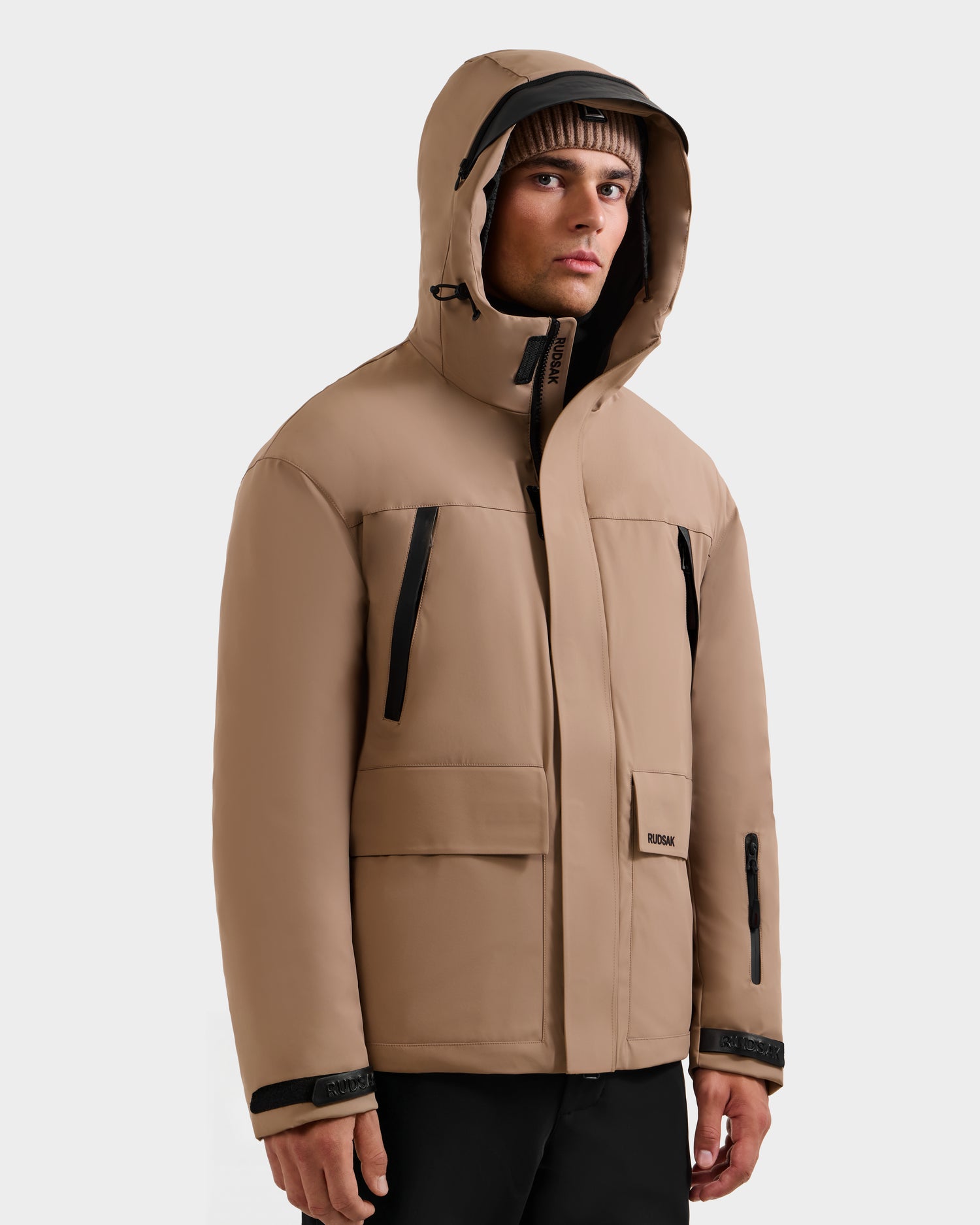 A man in the Rudsak ETHAN - 6125589 Sand jacket stands against a plain light background, wearing black pants and a brown knit beanie, looking slightly to the side.