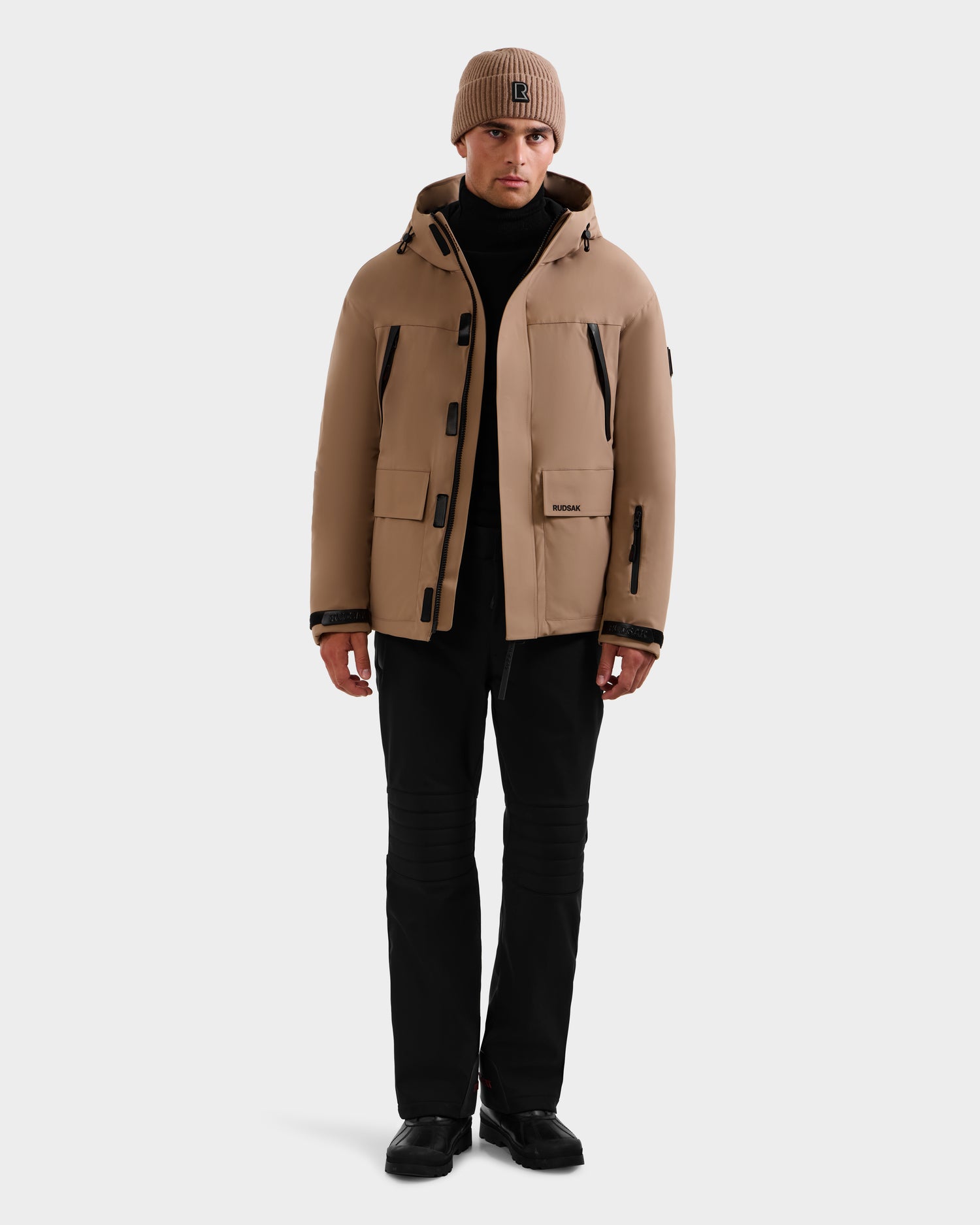 A person wearing the Rudsak ETHAN - 6125589 Sand men's ski jacket, a matching knit beanie, black turtleneck, pants, and shoes stands facing forward against a plain white background.
