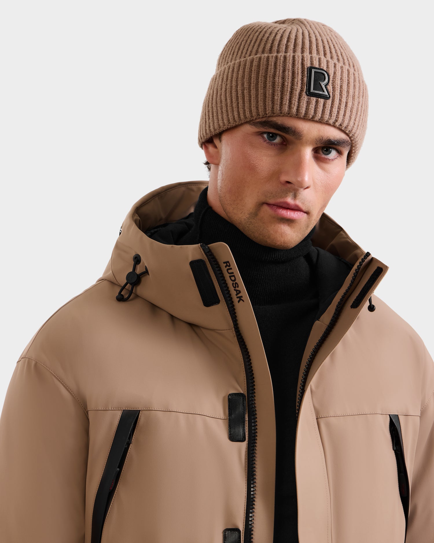 A man wearing the Rudsak ETHAN - 6125589 Sand men's ski jacket with black zippers over a black turtleneck and a ribbed beige beanie with a black R logo, looking at the camera. The snowboard jacket features RDS-certified down for warmth.