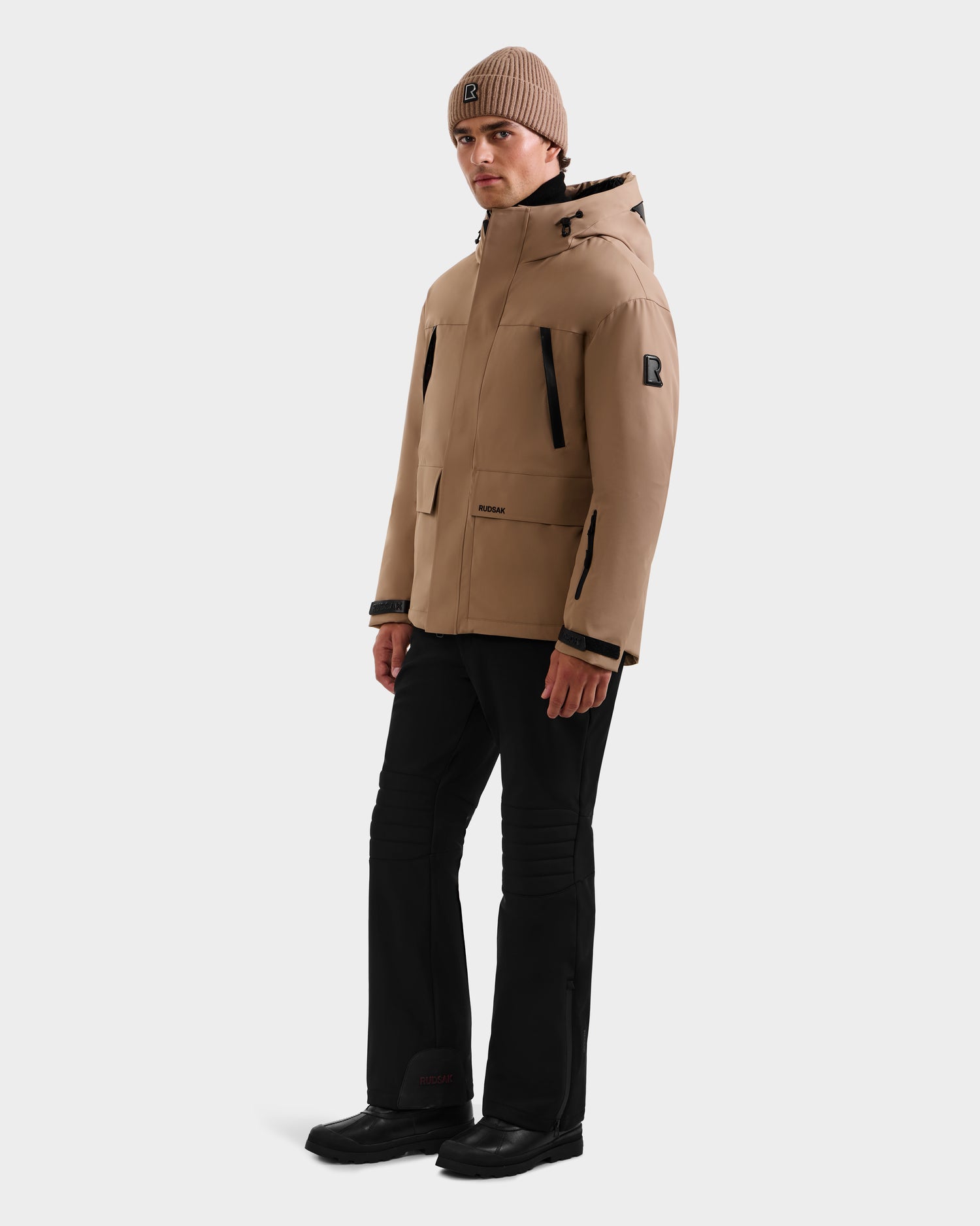A person wearing the Rudsak ETHAN - 6125589 Sand men’s ski jacket with RDS-certified down, a tan beanie, black pants, and black shoes stands against a plain white background, looking slightly to the side.