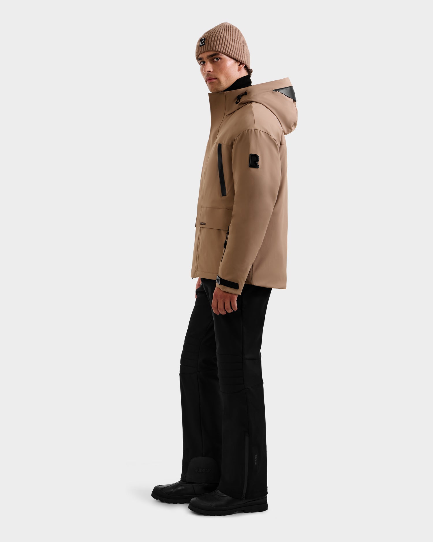 A person wearing the Rudsak ETHAN - 6125589 Sand jacket stands against a plain white background, paired with black pants, black shoes, and a tan beanie, looking over their shoulder.