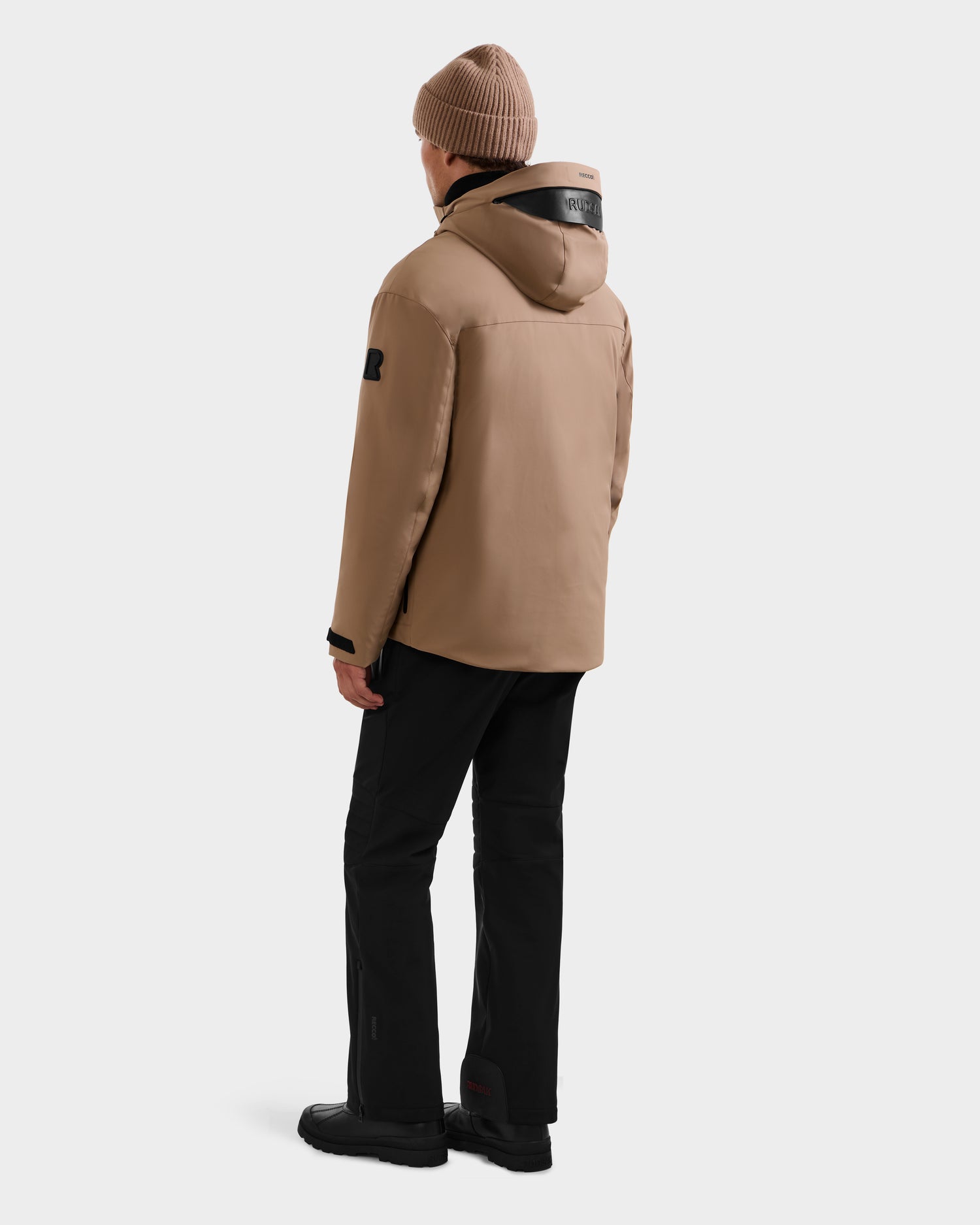 A person wearing the Rudsak ETHAN - 6125589 Sand men’s ski jacket, black pants, black shoes, and a brown knit beanie stands facing away against a plain light gray background.