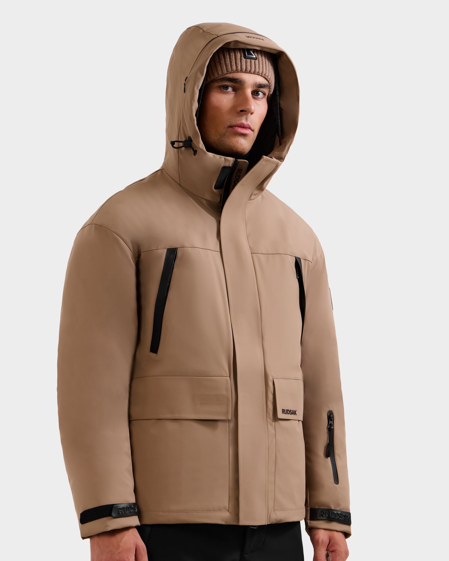A man wearing the Rudsak ETHAN - 6125589 Sand men’s ski jacket and a beige knit beanie stands against a plain light gray background.