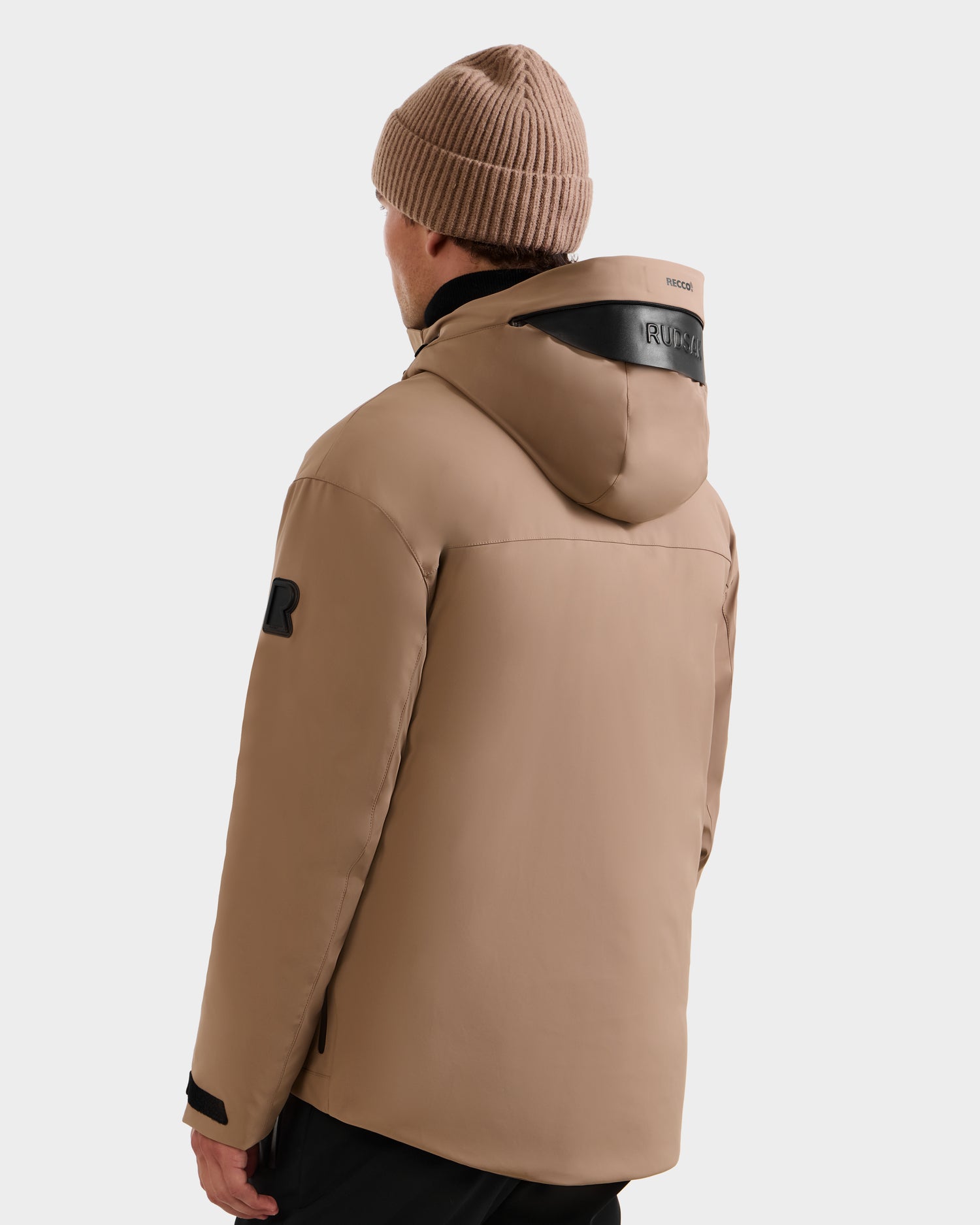 A person wearing the Rudsak ETHAN - 6125589 Sand men’s ski jacket with black accents and RDS-certified down fill, plus a beige knit beanie, faces away from the camera. The brand name appears on the hood’s back.