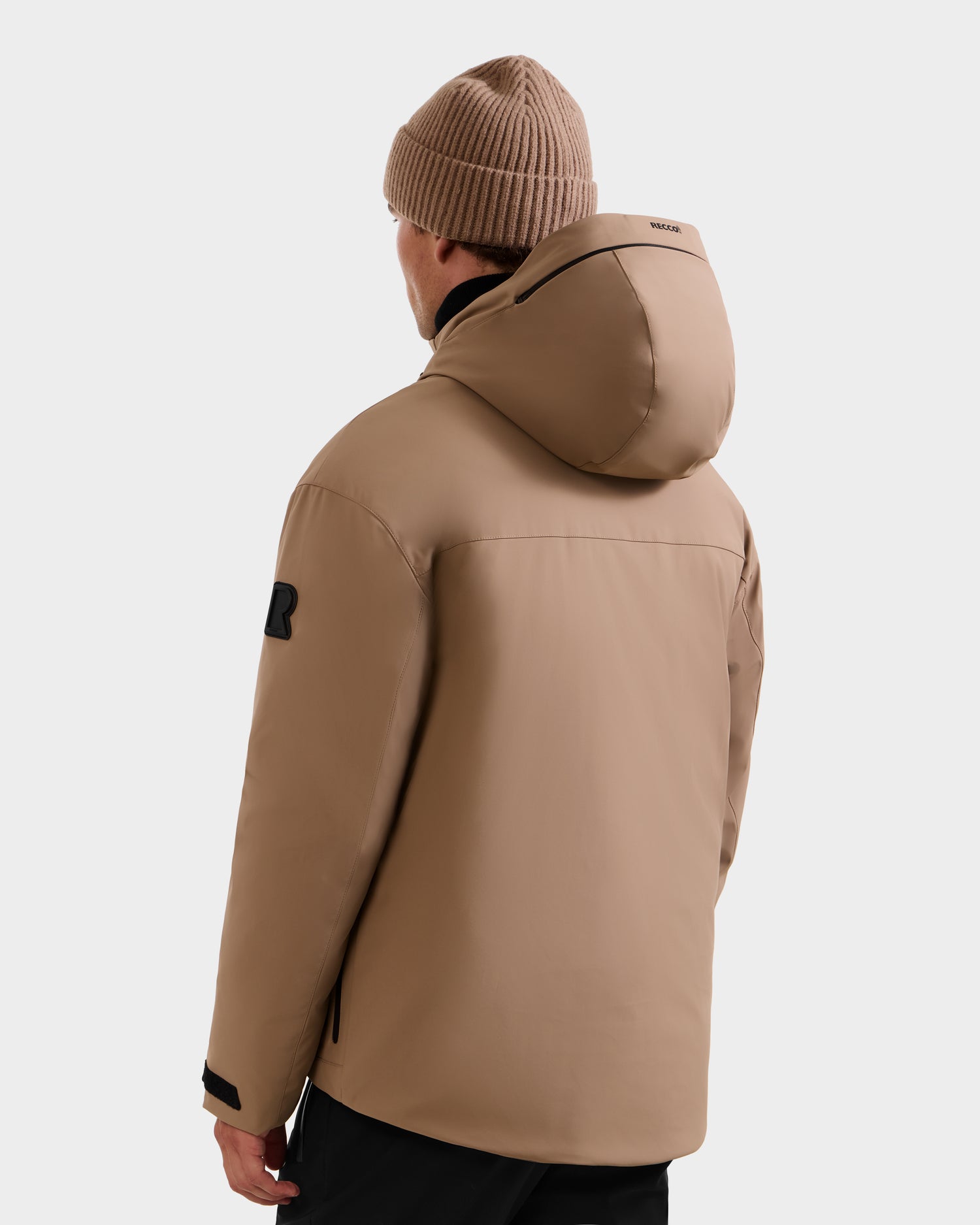 A person is shown from the back, facing slightly left, wearing Rudsak's ETHAN - 6125589 Sand men’s ski jacket with RDS-certified down, a matching knit beanie, black logo patch on the sleeve, and black zipper accents.