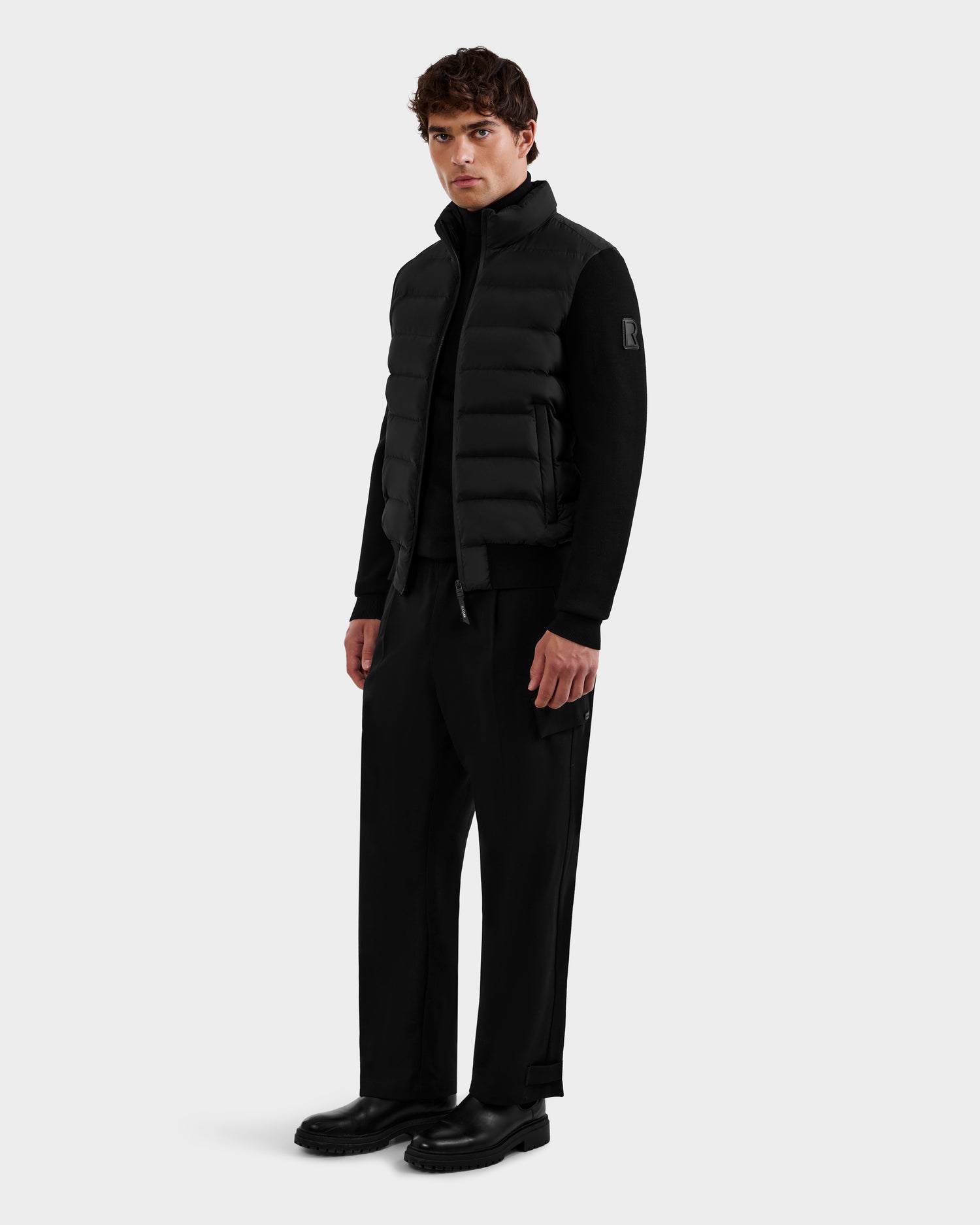 A man faces forward against a light gray background, wearing Rudsak's EUGENE - 6124721 Black men’s hybrid jacket as a puffer vest over a black zip-up shirt, paired with black wide-leg pants and shoes.