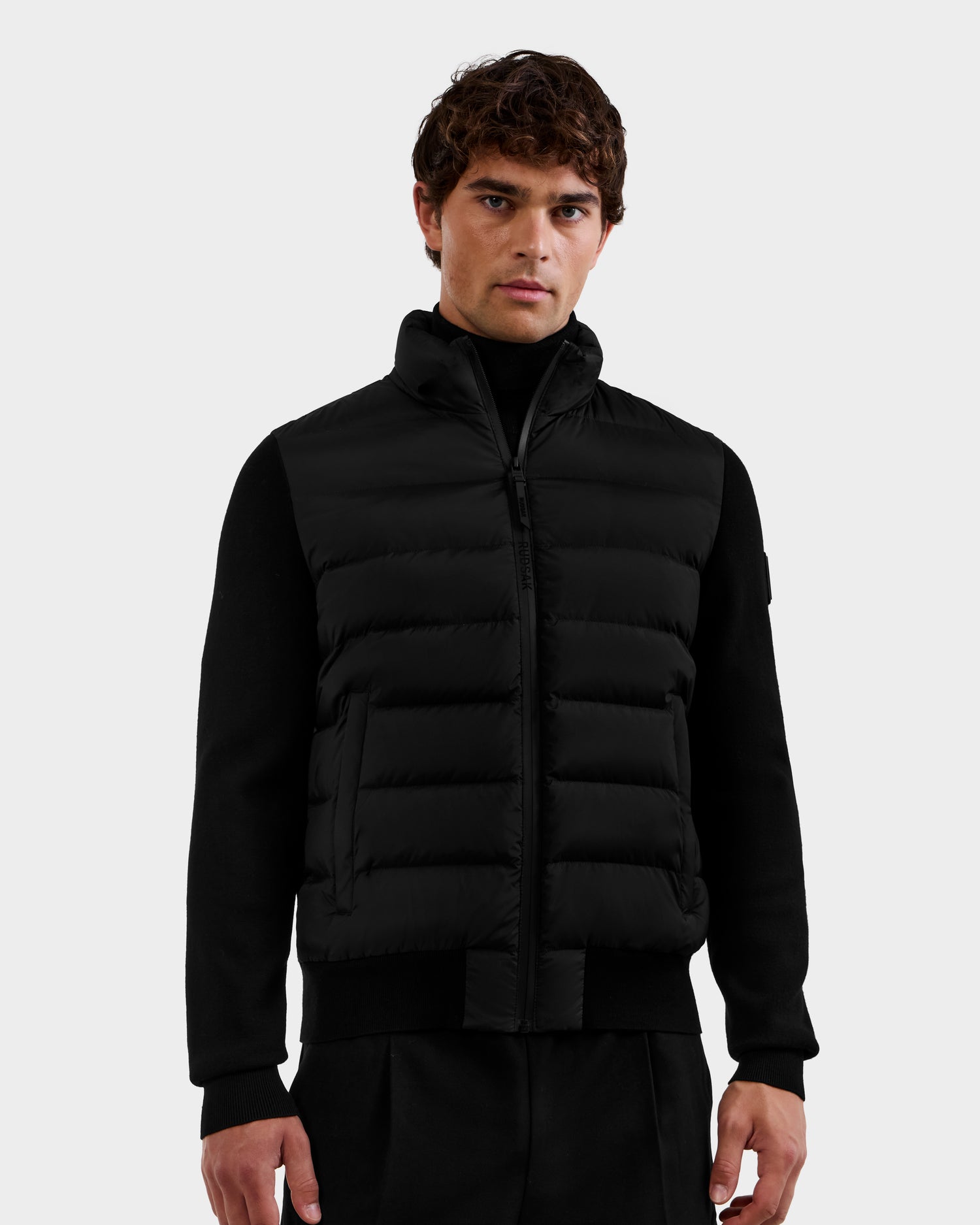 A man models the Rudsak EUGENE - 6124721 Black performance luxury jacket over black attire, standing against a plain light background and looking directly at the camera.