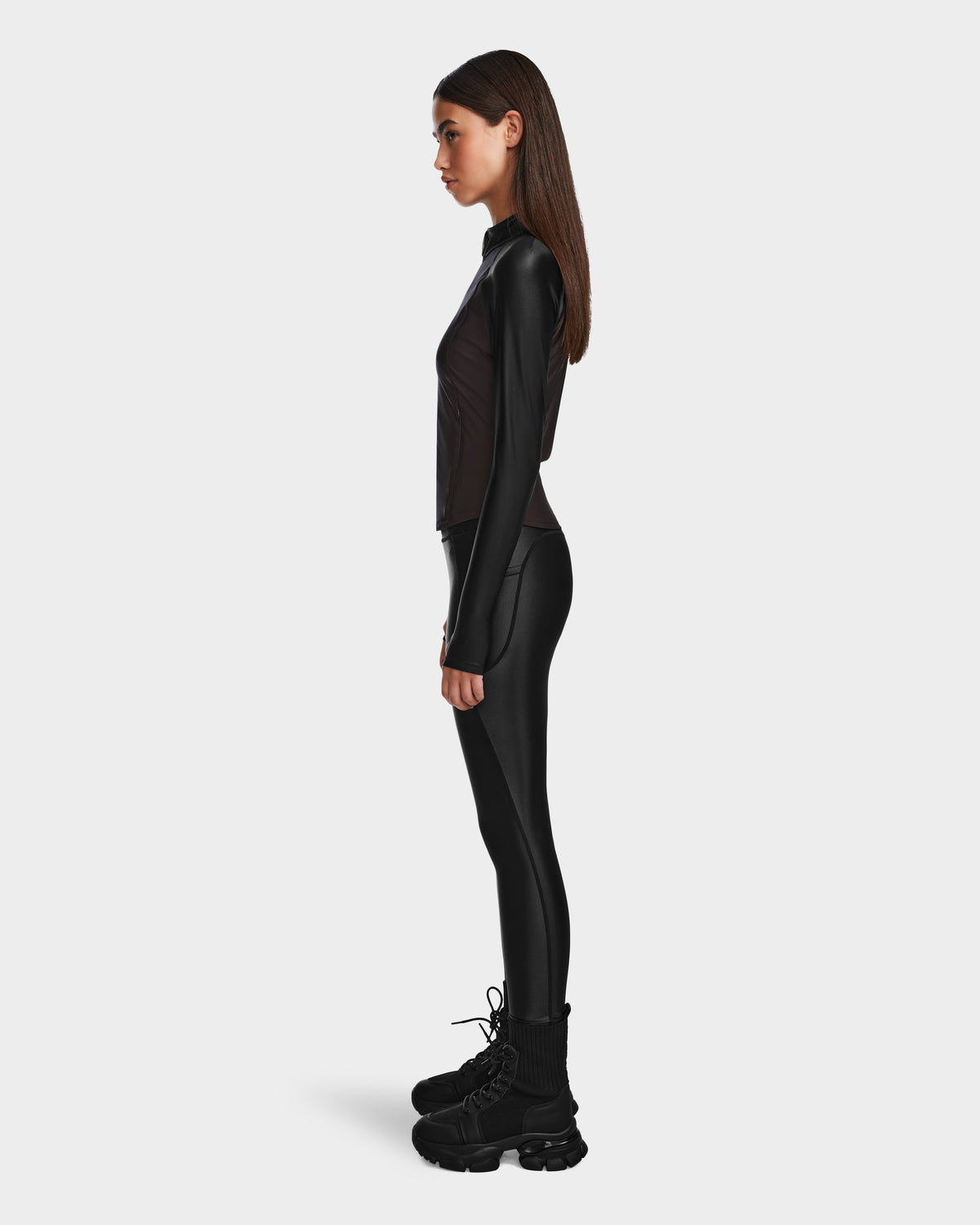 A woman stands in profile against a plain background, wearing the Rudsak EVA - 8524514 Black fitted zip-up top with waterproof zipper, matching black leggings, and chunky boots. Her hair is straight as she faces left.