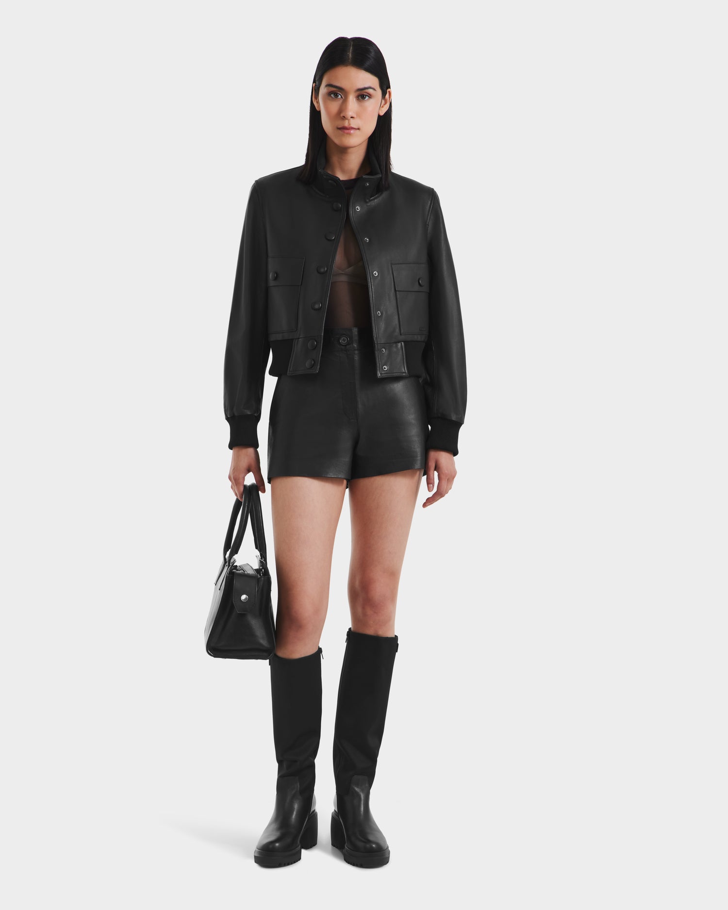 A woman with straight dark hair and a neutral expression faces forward in a Rudsak EVERLEIGH - 8125547 Black leather bomber jacket, black high-waisted shorts, knee-high boots, and holds a black handbag against a plain white background.