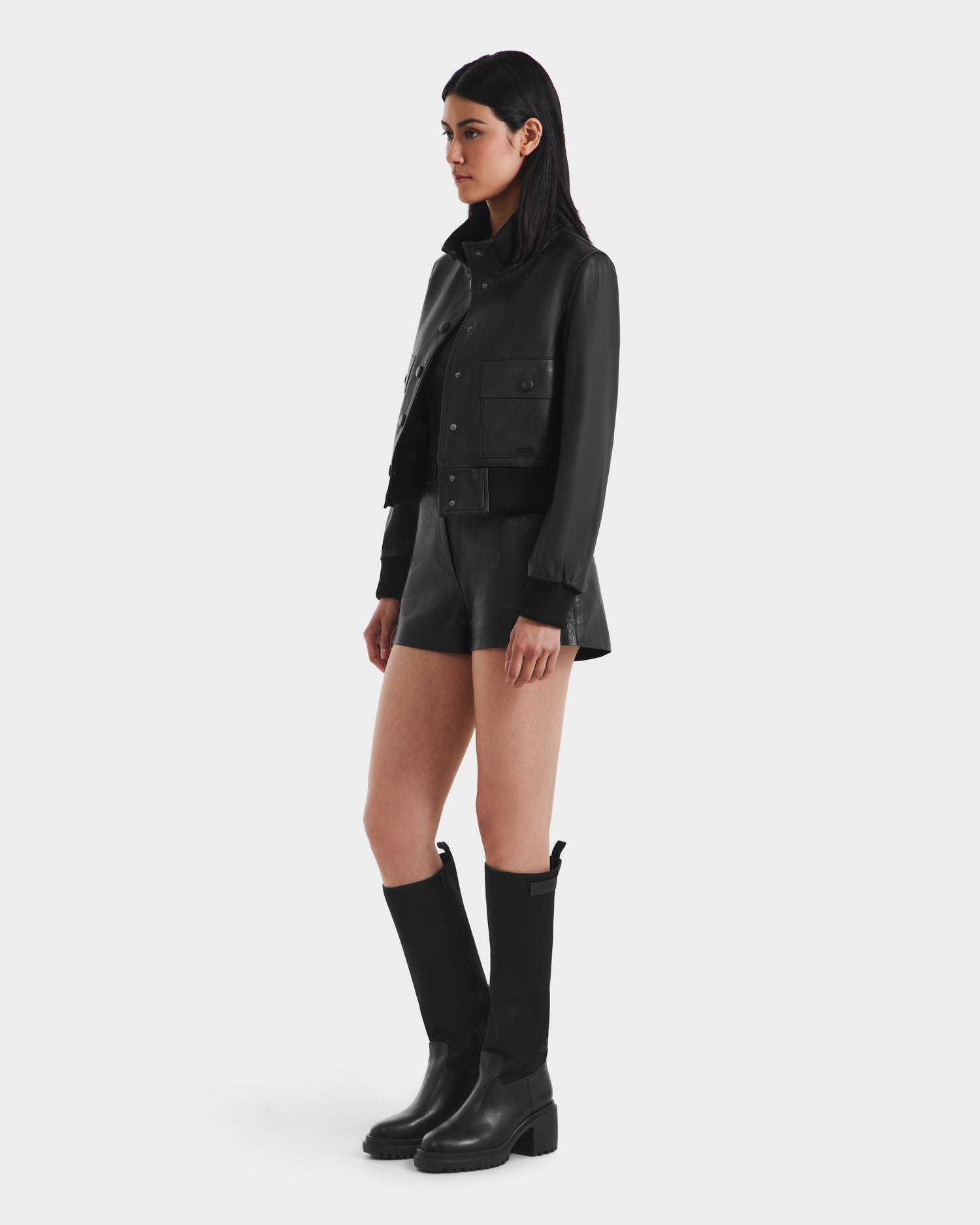 A woman poses against a plain white background wearing the Rudsak EVERLEIGH - 8125547 Black leather bomber jacket, paired with black shorts and knee-high boots, looking slightly to the side with her arms relaxed at her sides.