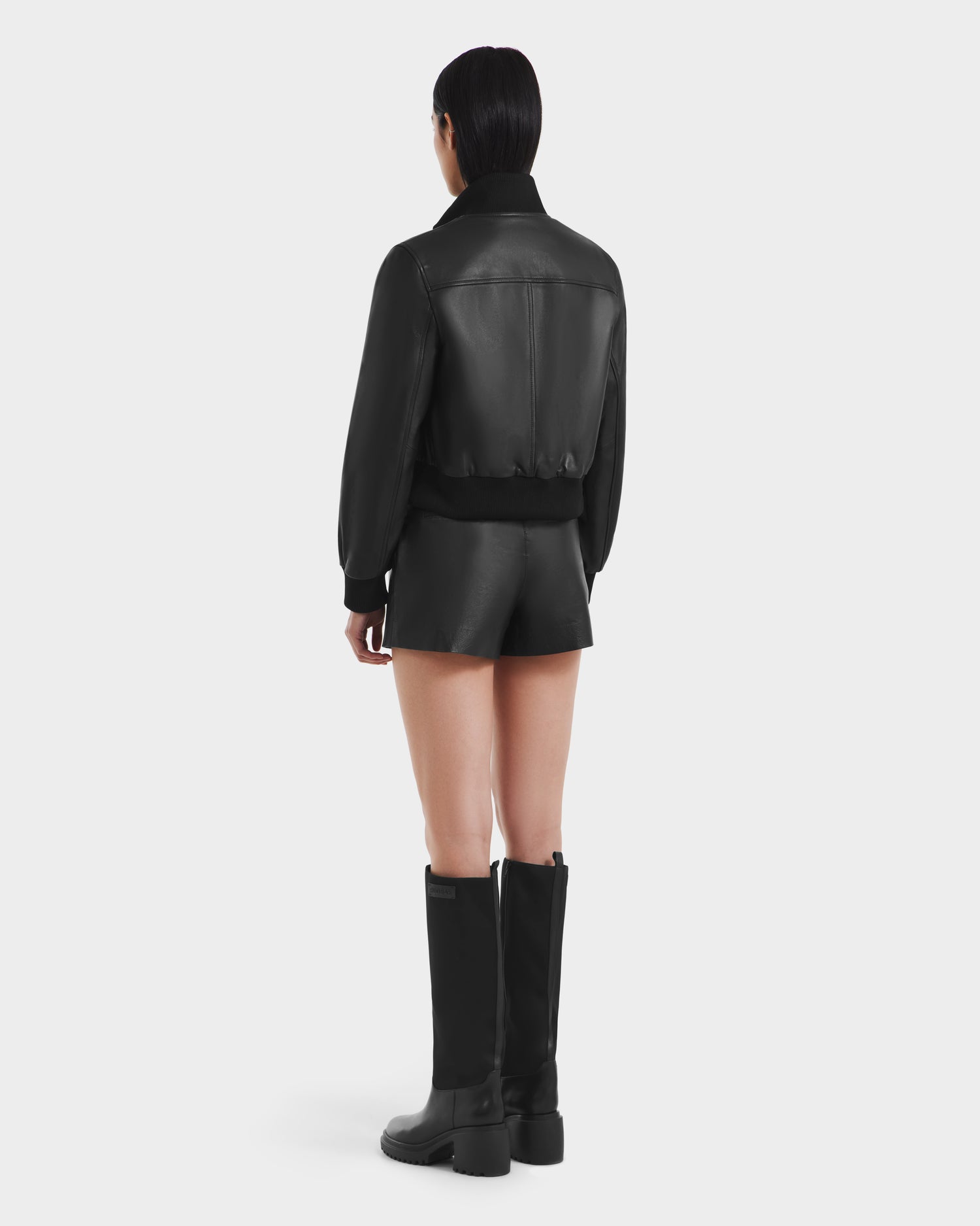A woman in a sleek Rudsak EVERLEIGH - 8125547 Black leather bomber jacket paired with shorts.