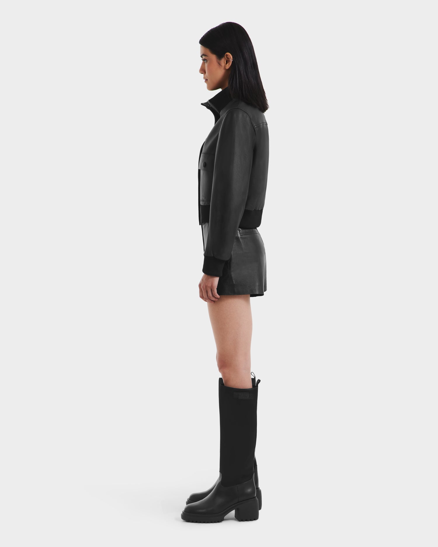 A woman in profile poses against a plain background, wearing the Rudsak EVERLEIGH - 8125547 Black women’s leather bomber jacket, paired with a black mini skirt and tall black boots.