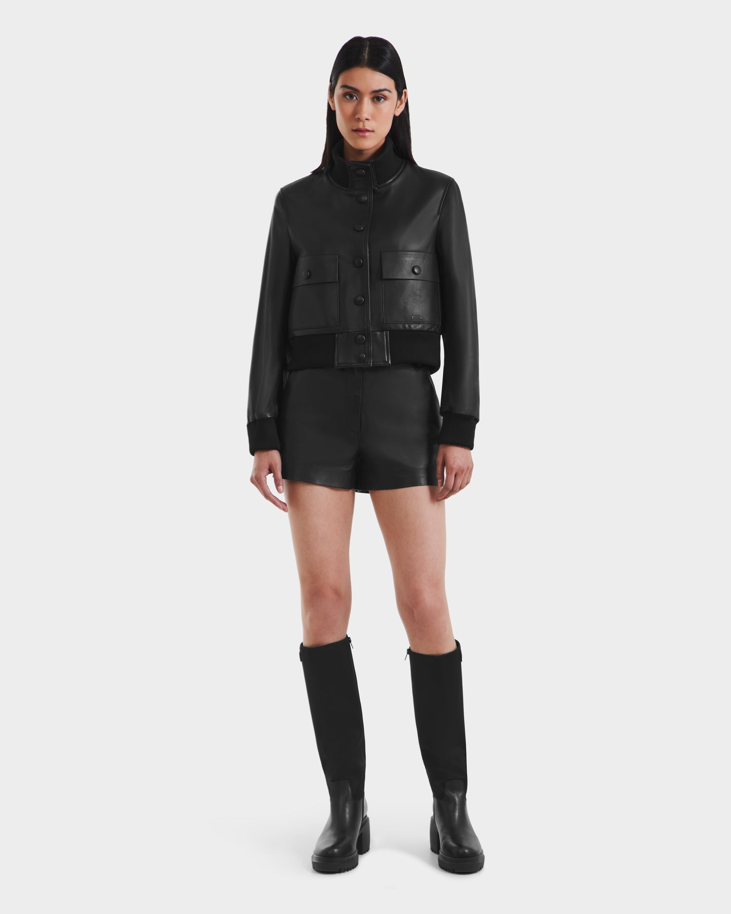 A woman faces forward against a plain white background, wearing the Rudsak EVERLEIGH - 8125547 Black women’s leather bomber jacket, paired with black leather shorts and knee-high black boots.