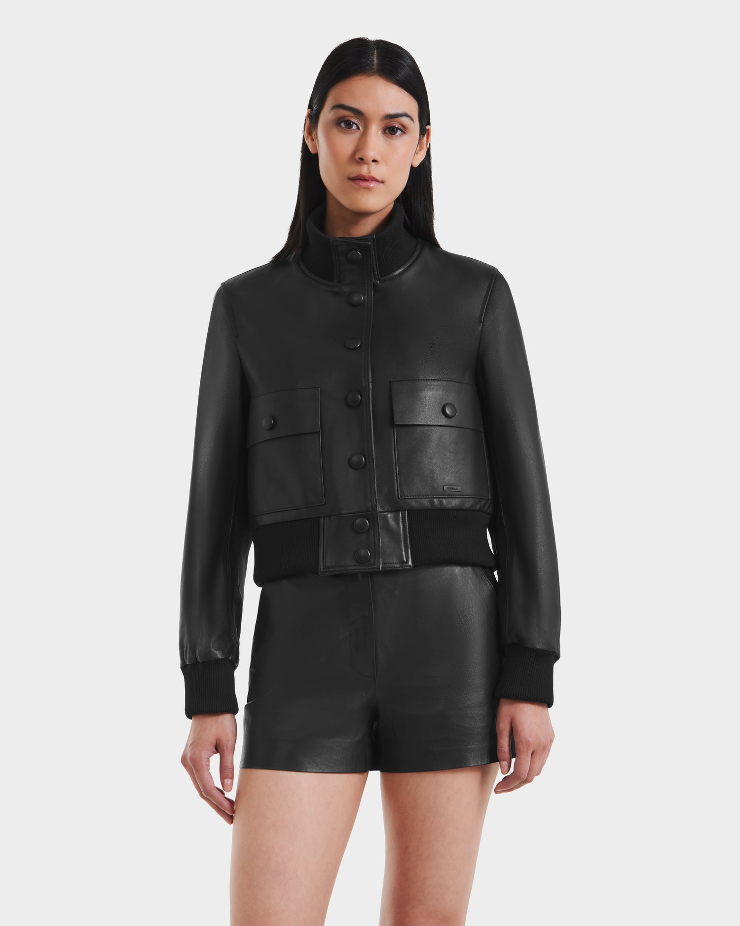 A woman faces forward, wearing the Rudsak EVERLEIGH - 8125547 Black leather bomber jacket with large pockets and matching black leather shorts, set against a plain white background.