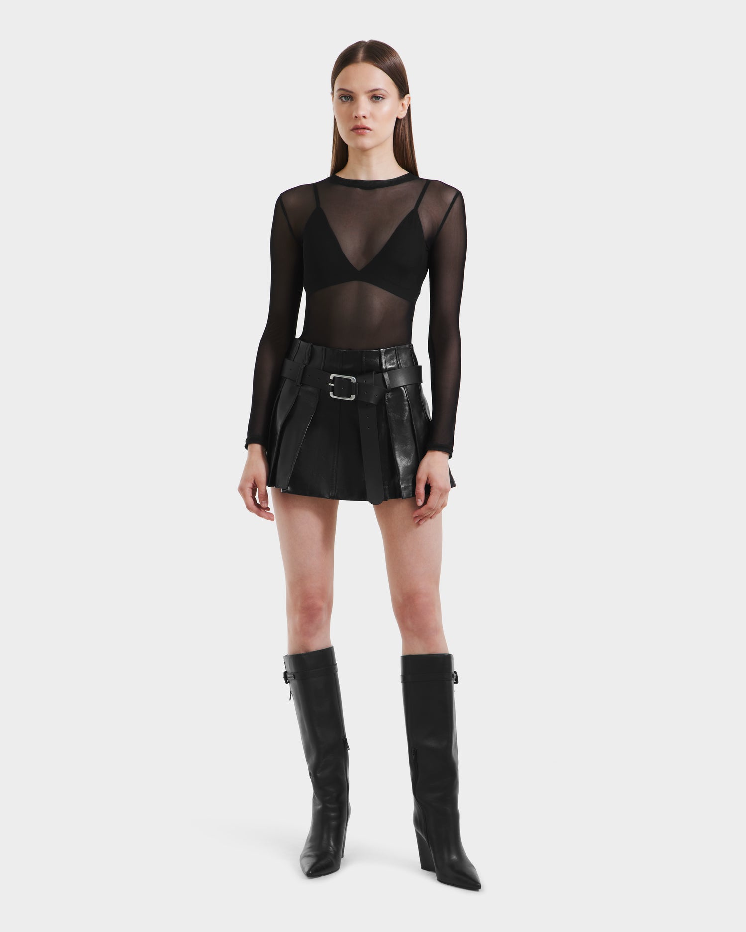 A woman with straight brown hair and a neutral expression faces forward, wearing the Rudsak FELICITY - 8525650 Black: a sheer long-sleeve top over a black bra, a high-waisted leather skirt with belt details, and knee-high black boots.
