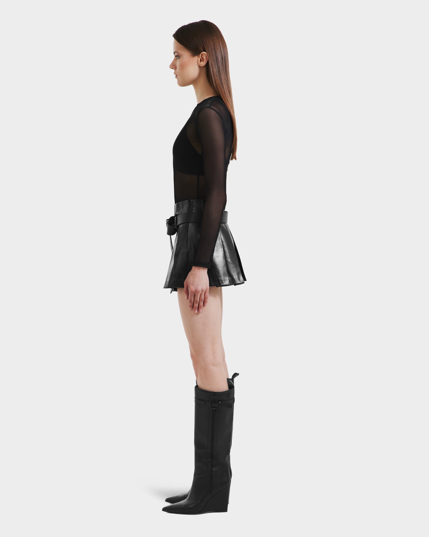 A woman with straight brown hair stands in profile against a white background, wearing a sheer black top, knee-high black boots, and the FELICITY - 8525650 Black leather mini skirt by Rudsak.