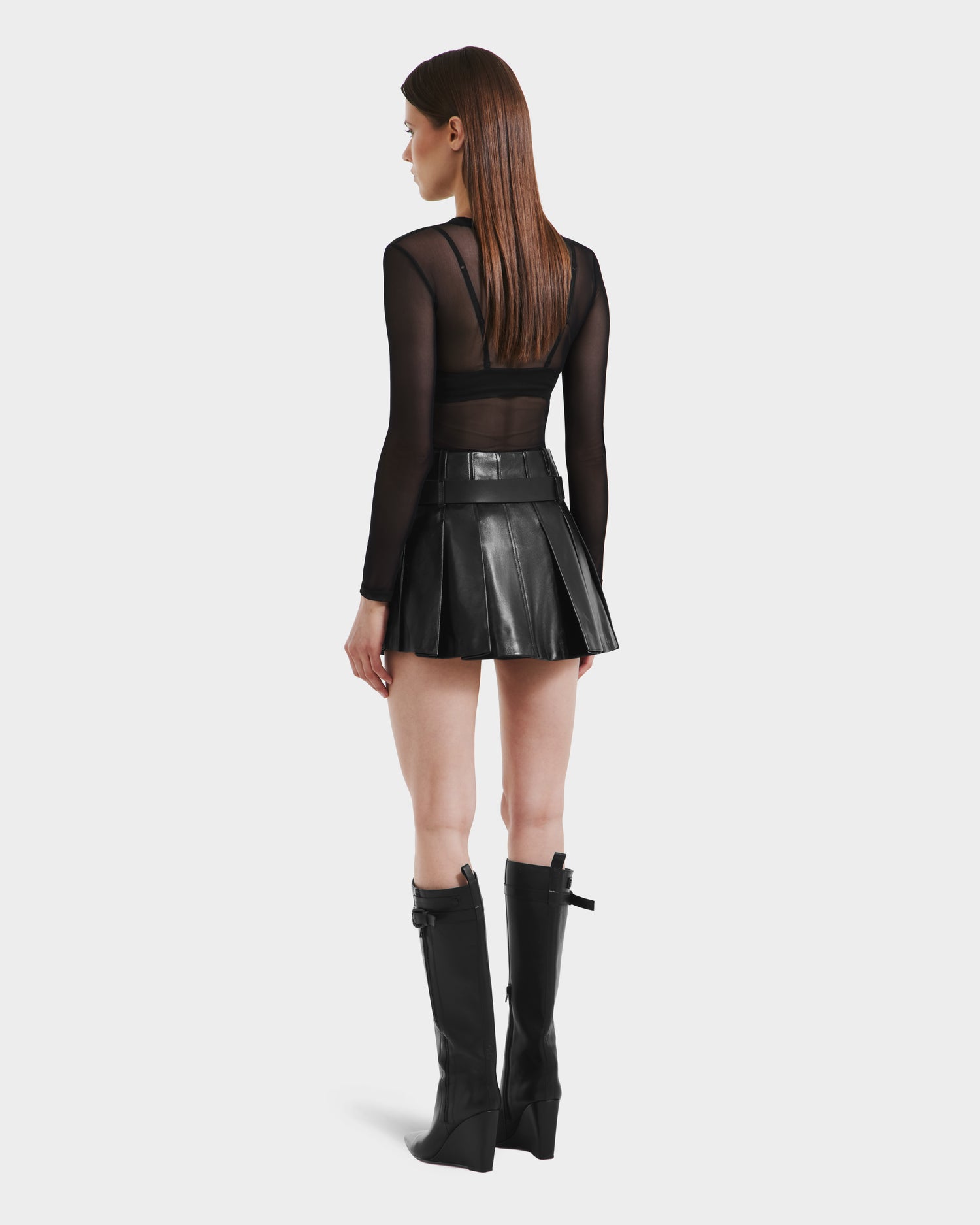 A woman stands facing away against a white background, wearing the Rudsak FELICITY - 8525650 Black sheer long-sleeve top, a high-waisted pleated leather skirt, and tall black wedge-heeled boots.