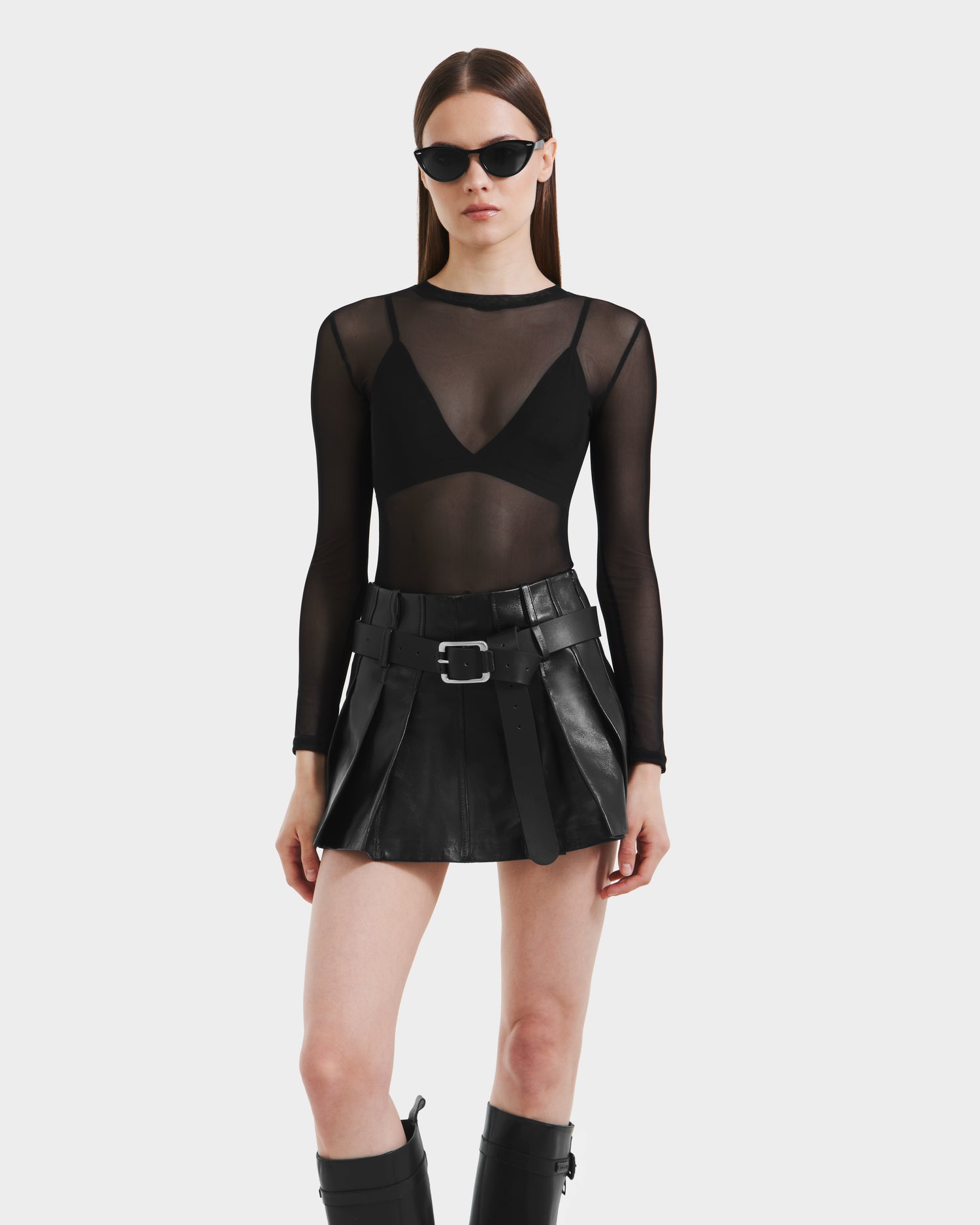 A woman stands against a plain white background wearing Rudsak’s FELICITY - 8525650 Black belted leather mini skirt, black knee-high boots, a sheer long-sleeved top over a black bra, and black sunglasses.