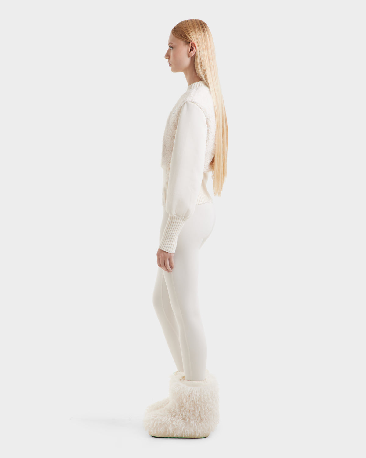 A woman with long straight blonde hair stands in profile, wearing Rudsak’s FLORENCE - 8525546 Iceland, a cozy-glam look with a white sweater, leggings, and fluffy faux fur womens boots against a plain light backdrop.