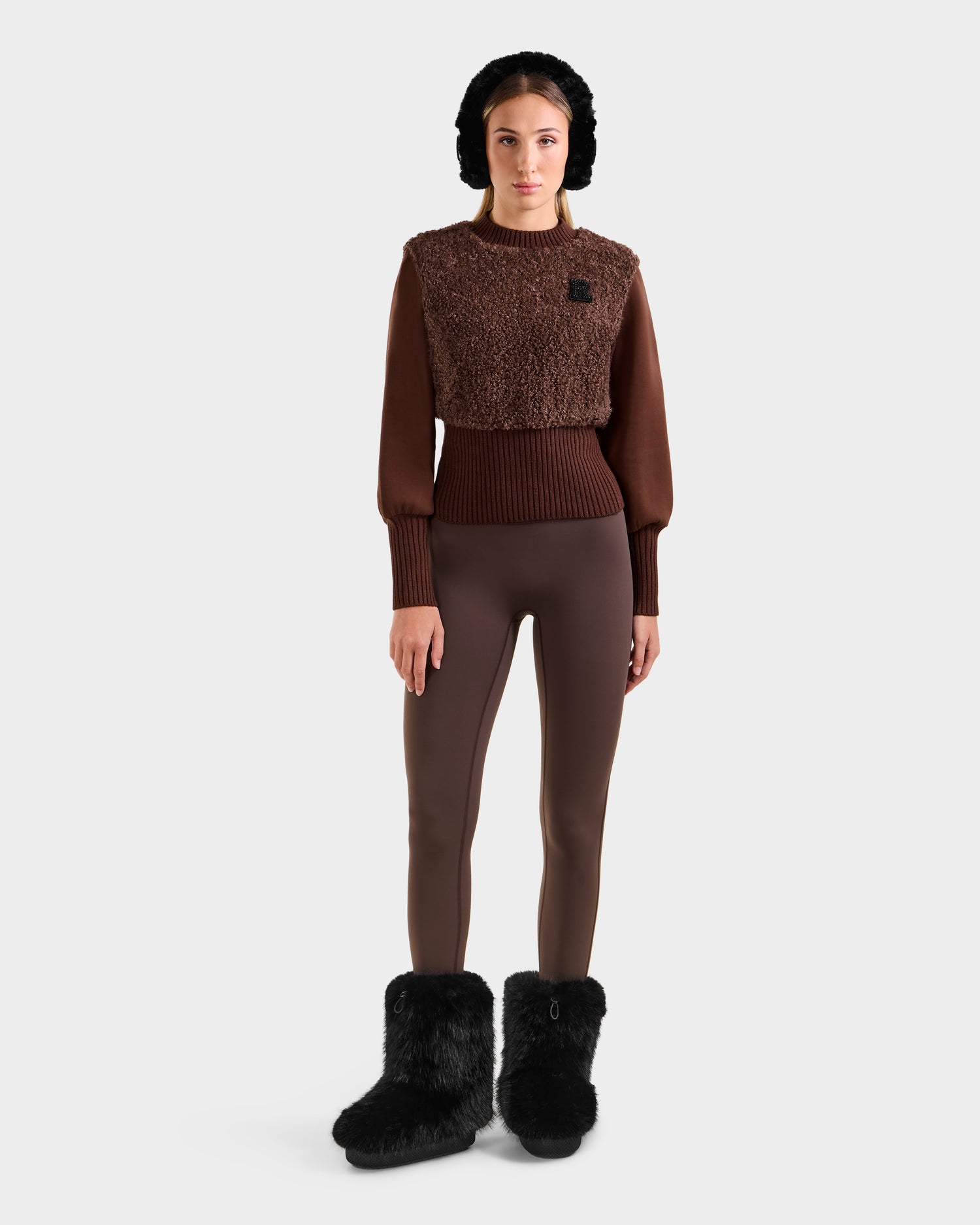 A woman wears the Rudsak FLORENCE - 8525546 Mocha cropped top, paired with brown leggings, black furry boots, and a black fur hat—an après-ski essential—standing forward against a plain light background.